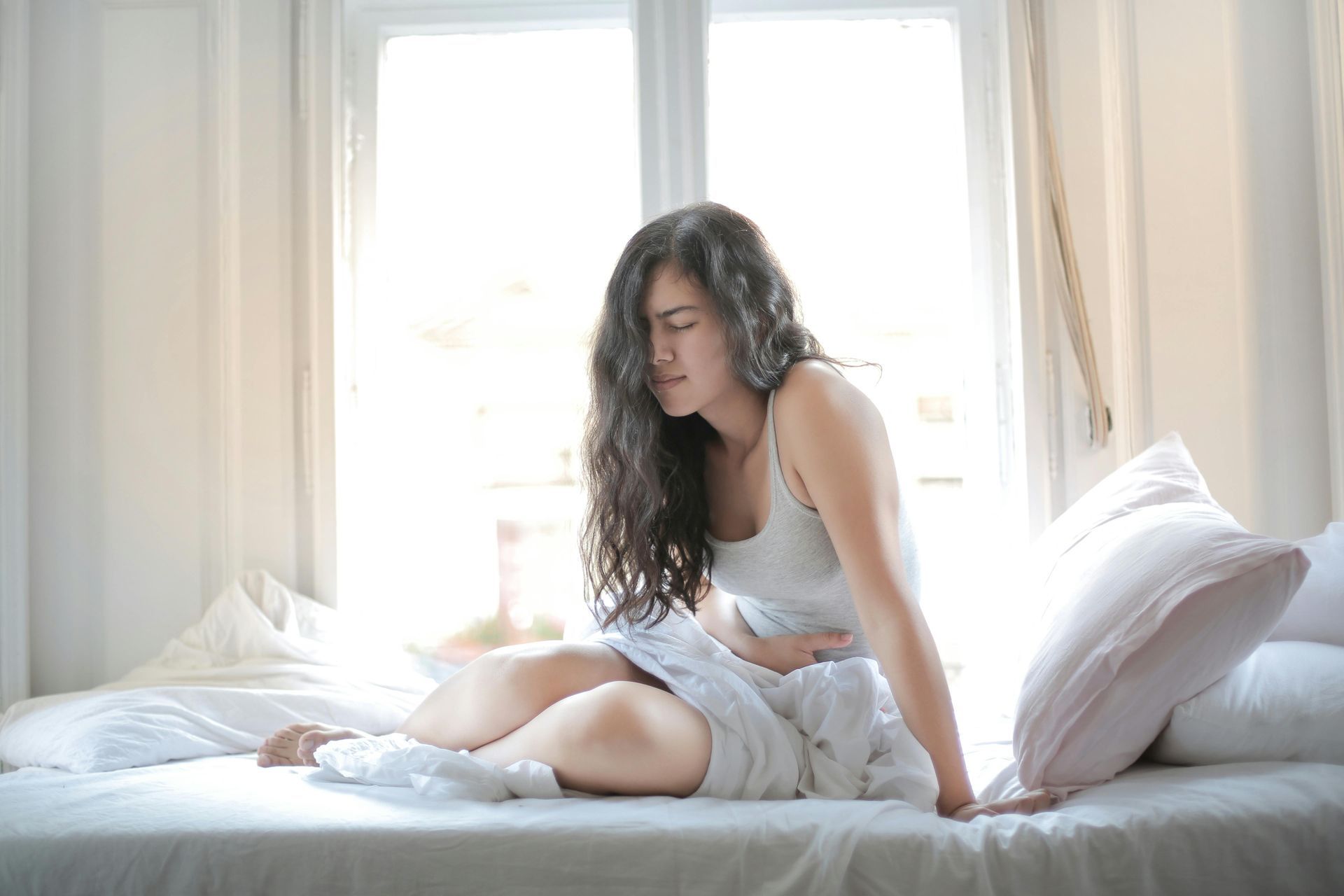 Woman with stomach pain, sitting on bed, near window, holding stomach, looking pained.