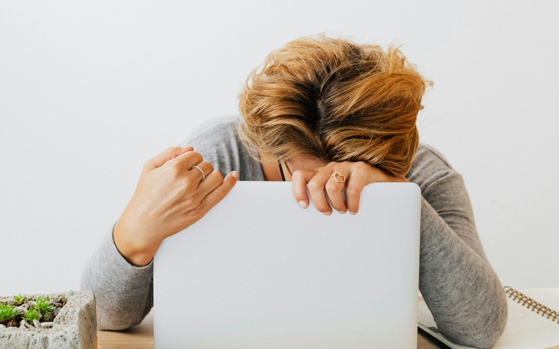 A person in a gray sweater sits at a wooden desk with their head resting on a closed laptop, appearing overwhelmed.