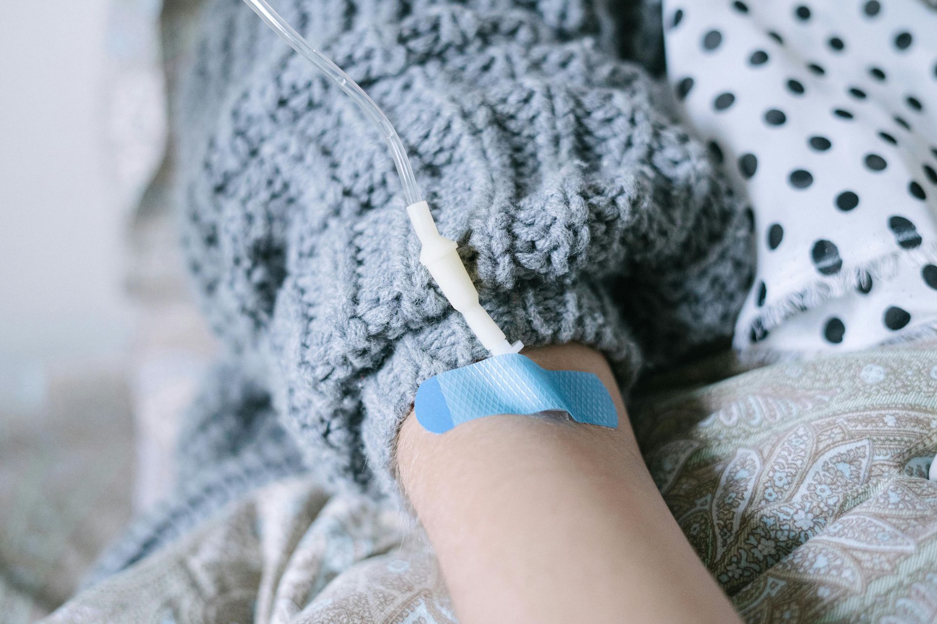 Person's arm with IV needle and blue medical tape. Wearing gray sweater.