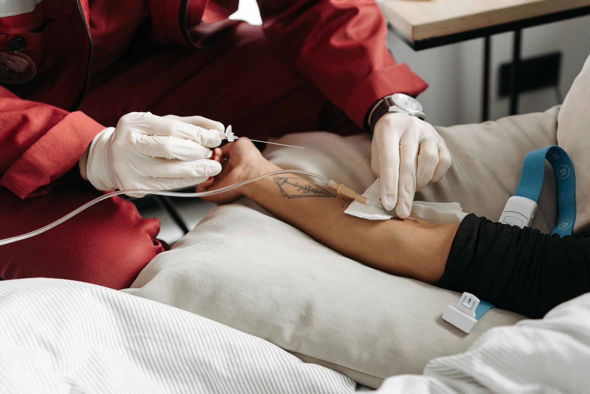 Person in red jacket inserting IV into patient's arm on a bed. White-gloved hands, blue strap.