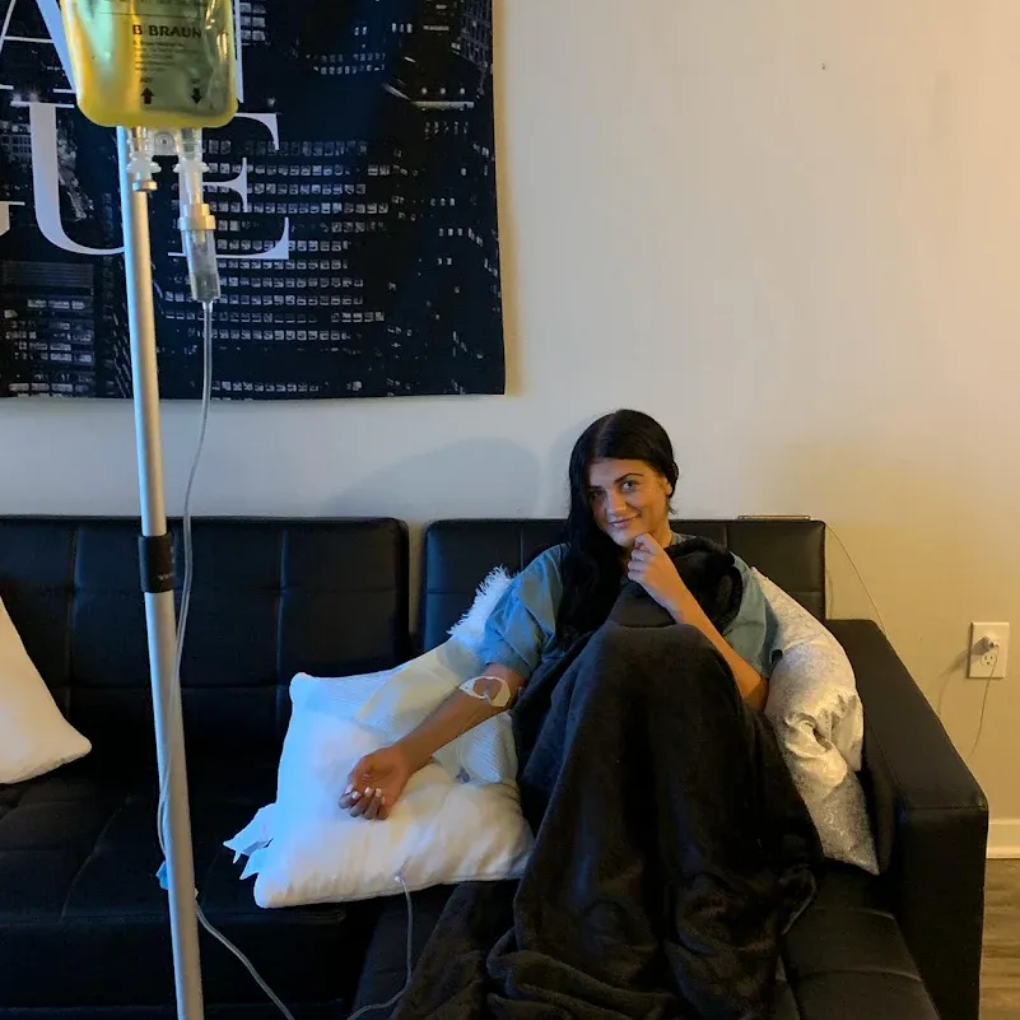 Woman receiving IV infusion, sitting on a black couch. Smiling, she is wrapped in a blanket in a room with artwork.