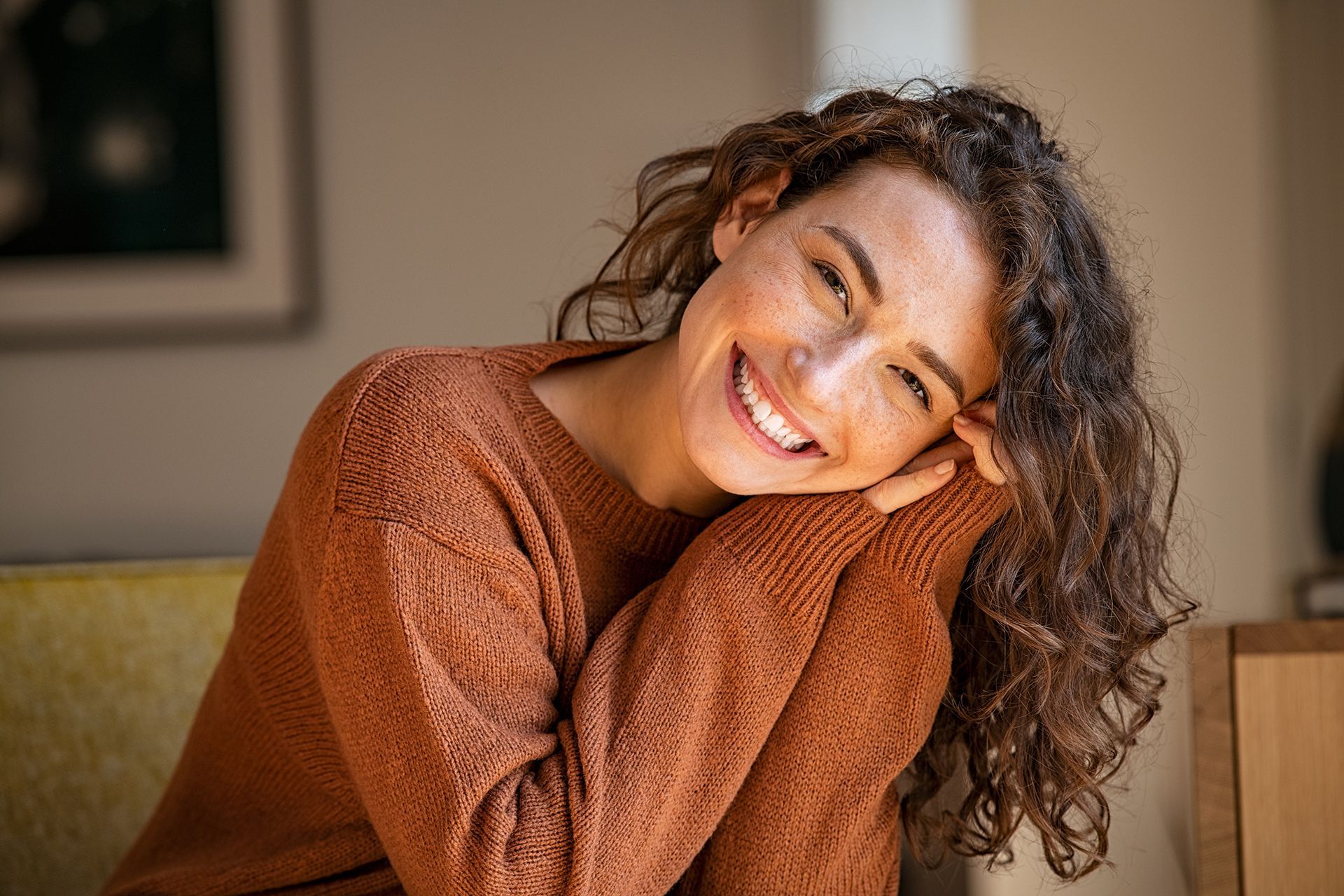 A smiling person with curly hair, wearing a brown sweater, rests their chin on their hands in a bright, indoor setting.