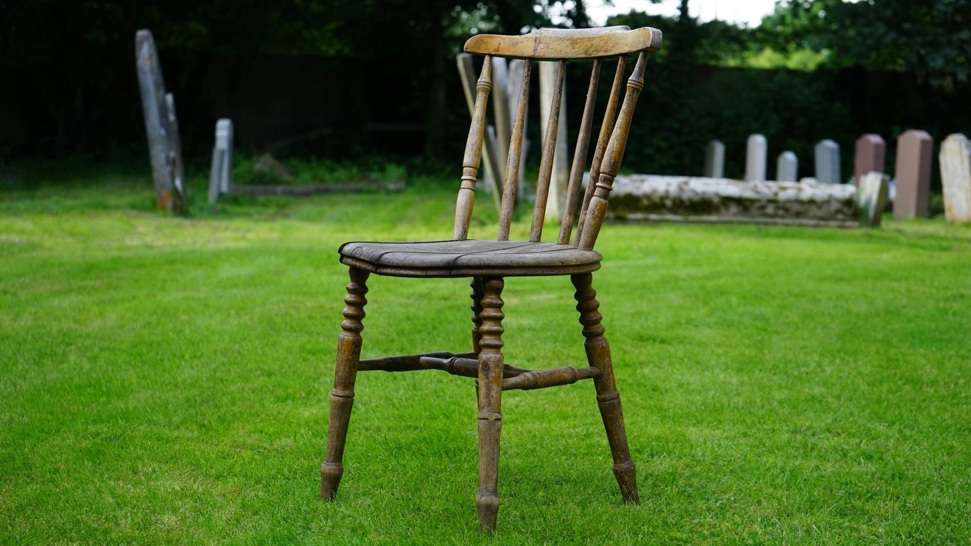 Worn wooden chair on green grass with headstones in the background.
