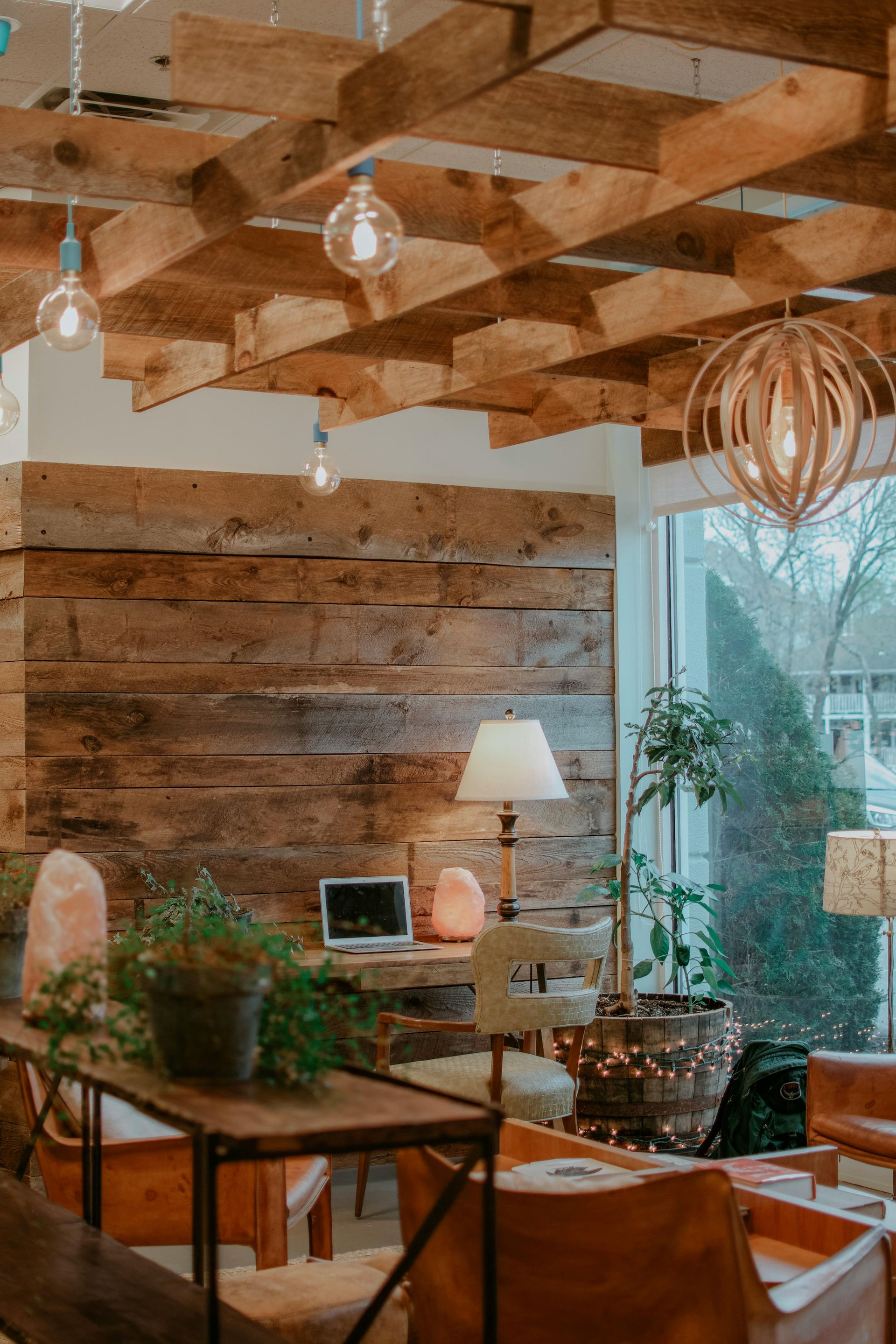 Cozy interior: wooden plank walls and ceiling, pendant lights, desk with laptop, plants, and natural light.