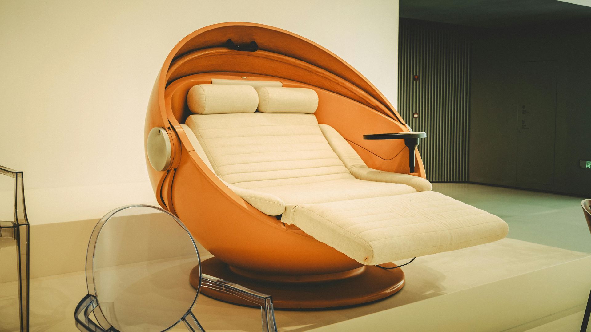 Orange egg-shaped lounge chair with white cushions. Brown base and side table, set against a neutral wall.