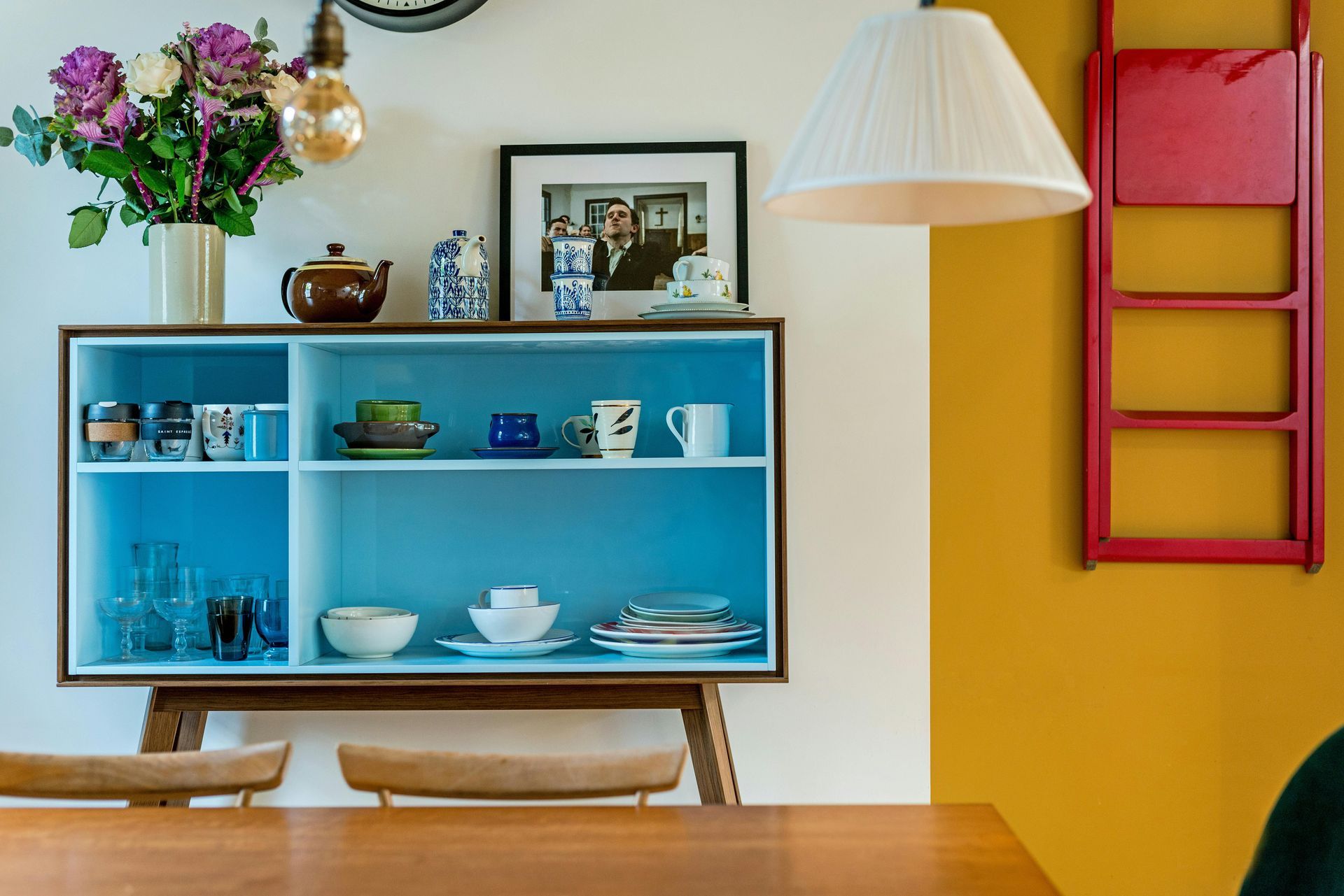 A dining area with a blue cabinet holding dishes, a red wall-mounted ladder, and yellow and white walls.