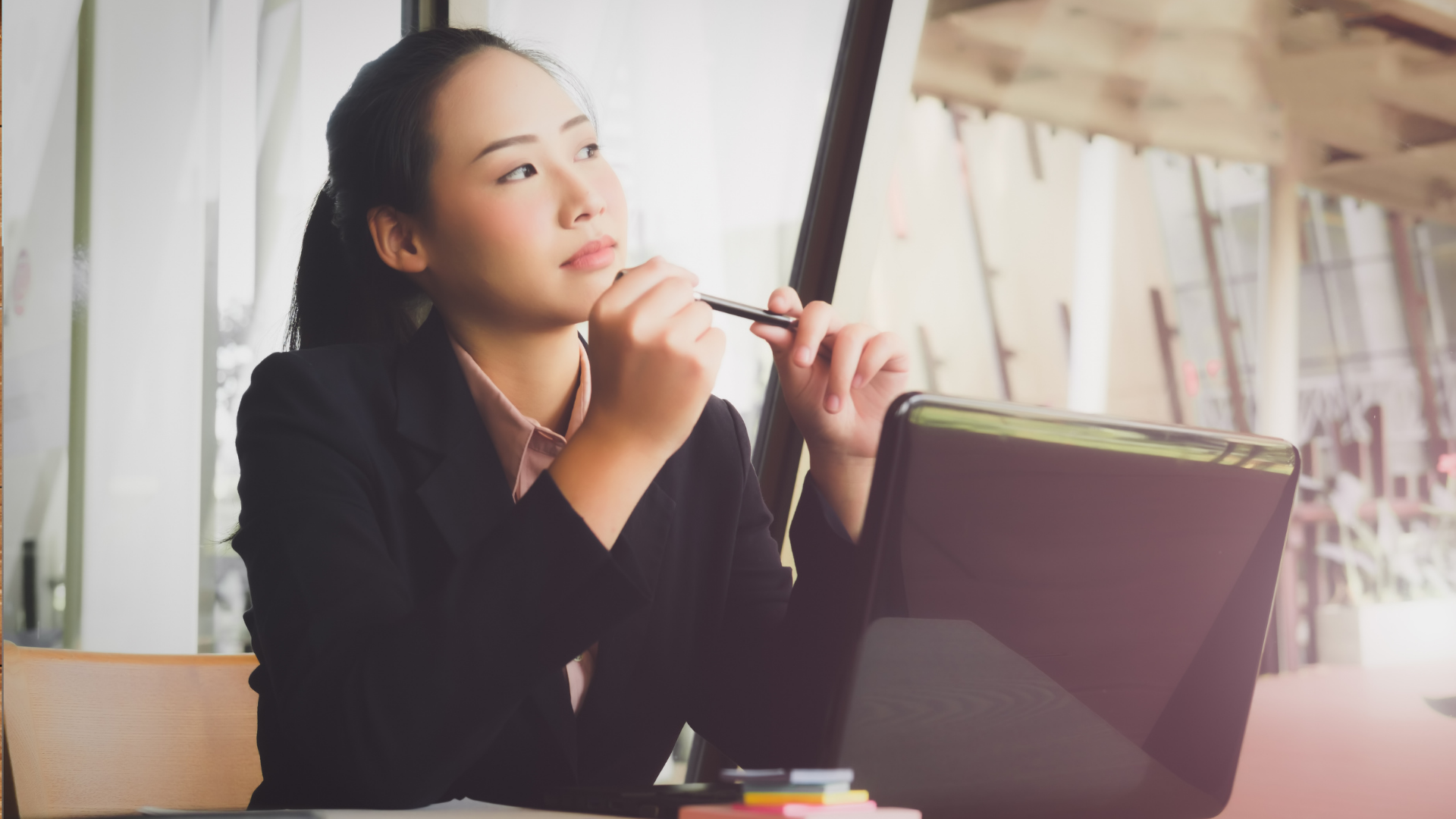 A woman is sitting at a desk with a laptop and a pen in her hand.