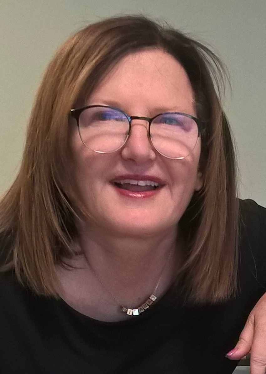 Smiling person with glasses and shoulder-length brown hair against a plain wall