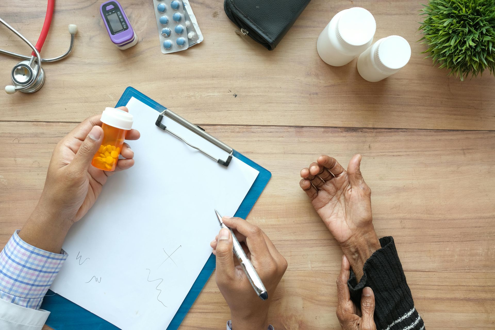 Hands handling medicine bottles and a clipboard on a wooden desk with assorted pills nearby