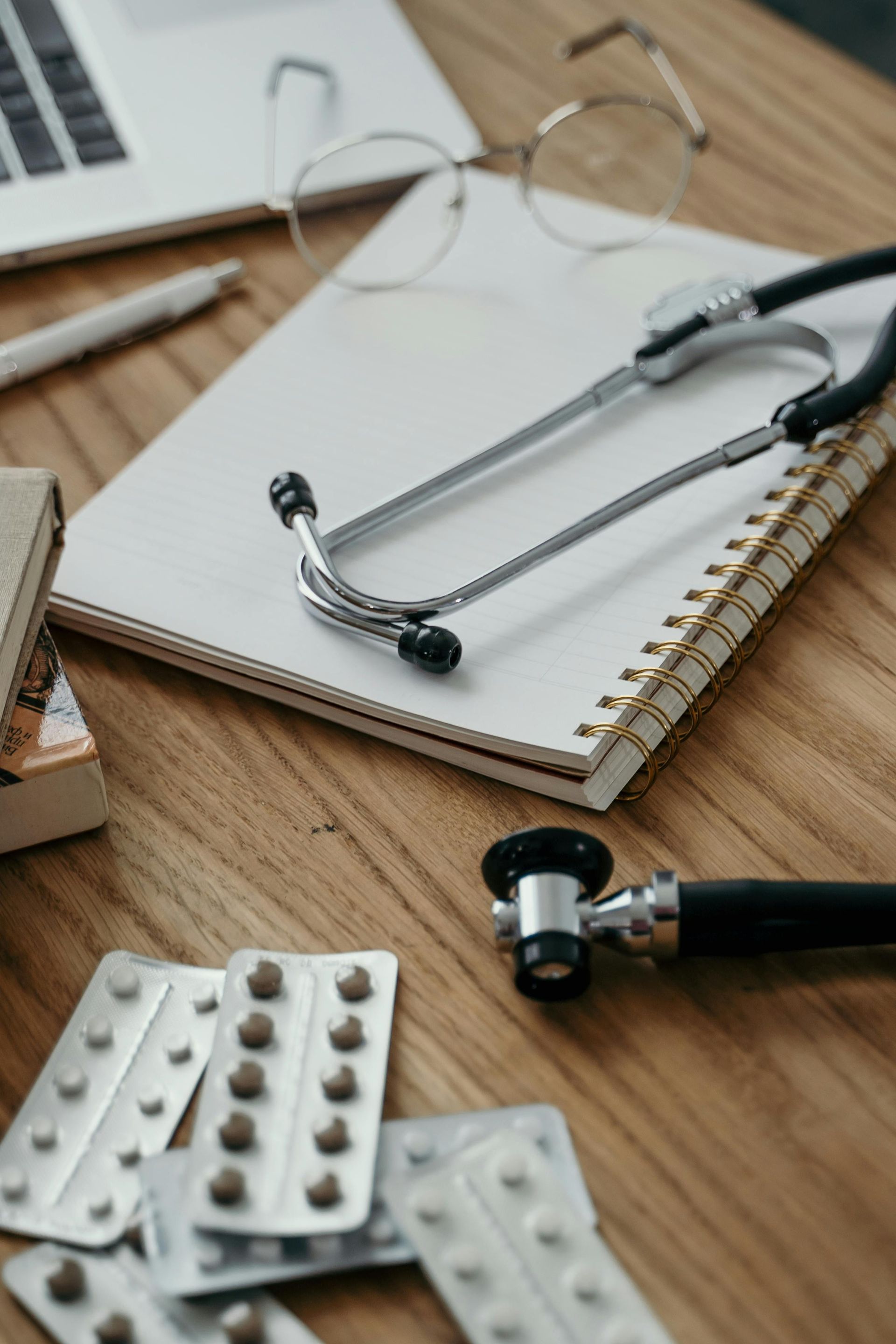 Stethoscope on a notebook beside pill blister packs and a laptop on a wooden desk