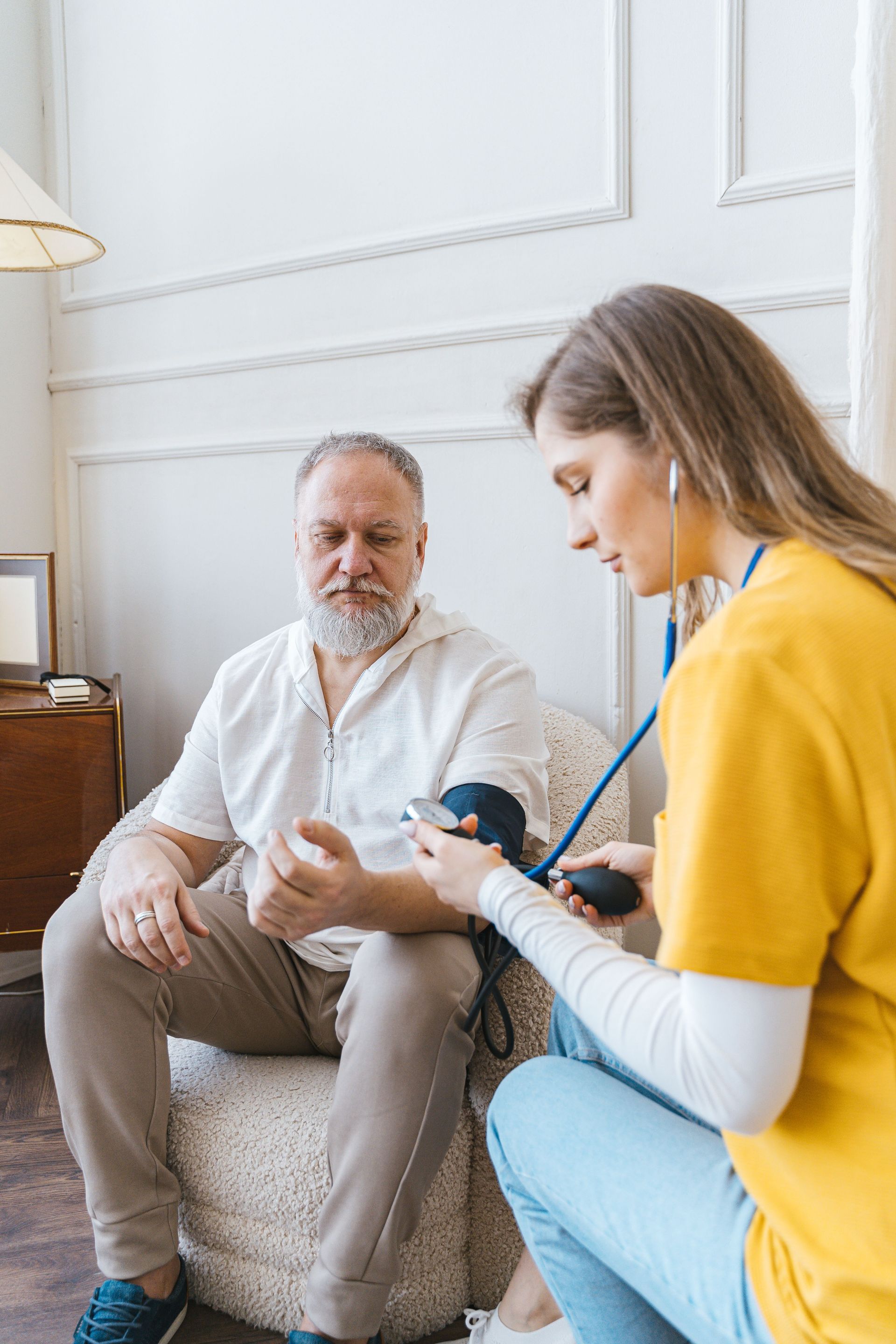 Woman kneels beside seated man, using a blood pressure cuff in a bright living room.