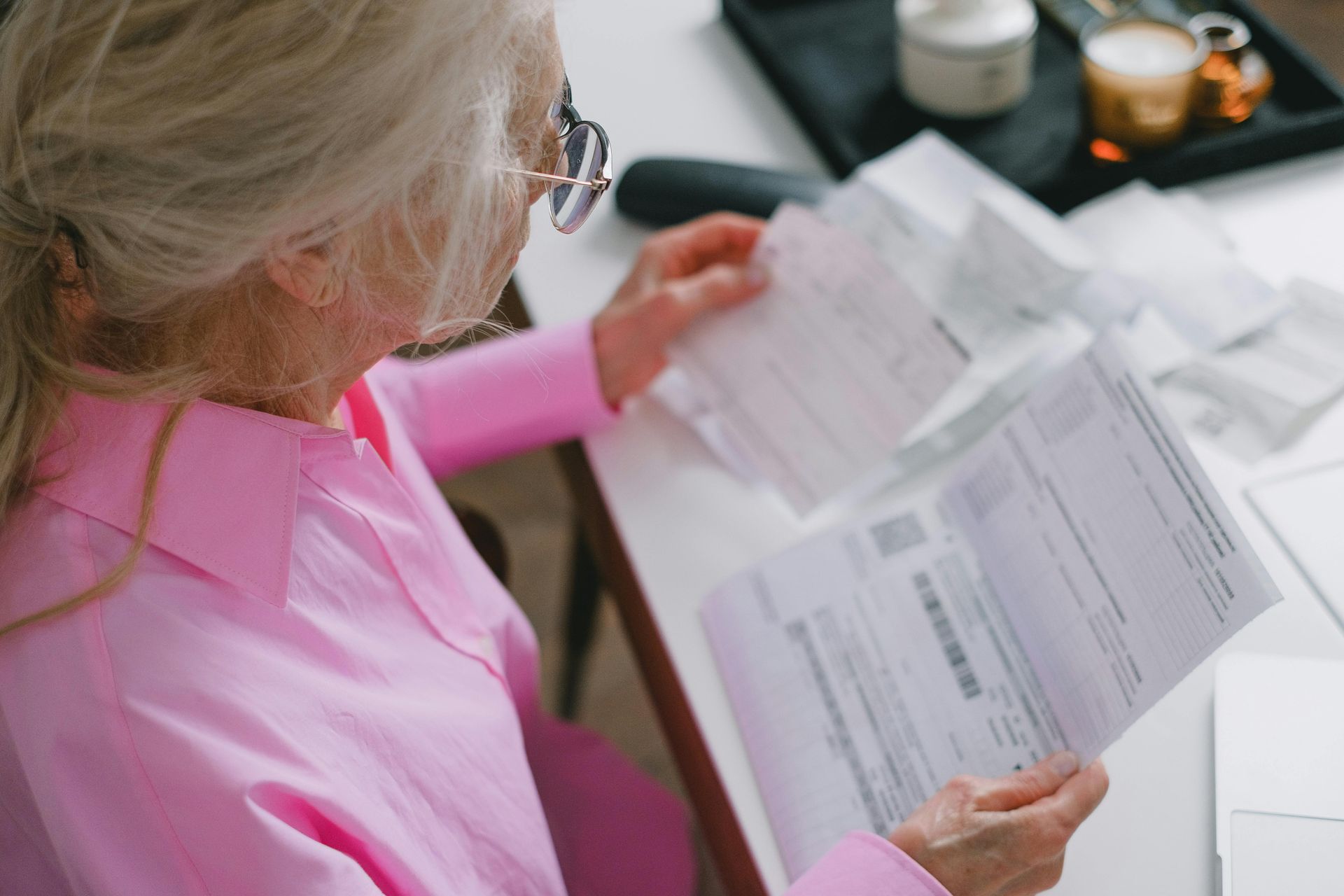 Person in a pink shirt reviewing paperwork at a desk with a laptop and coffee mug