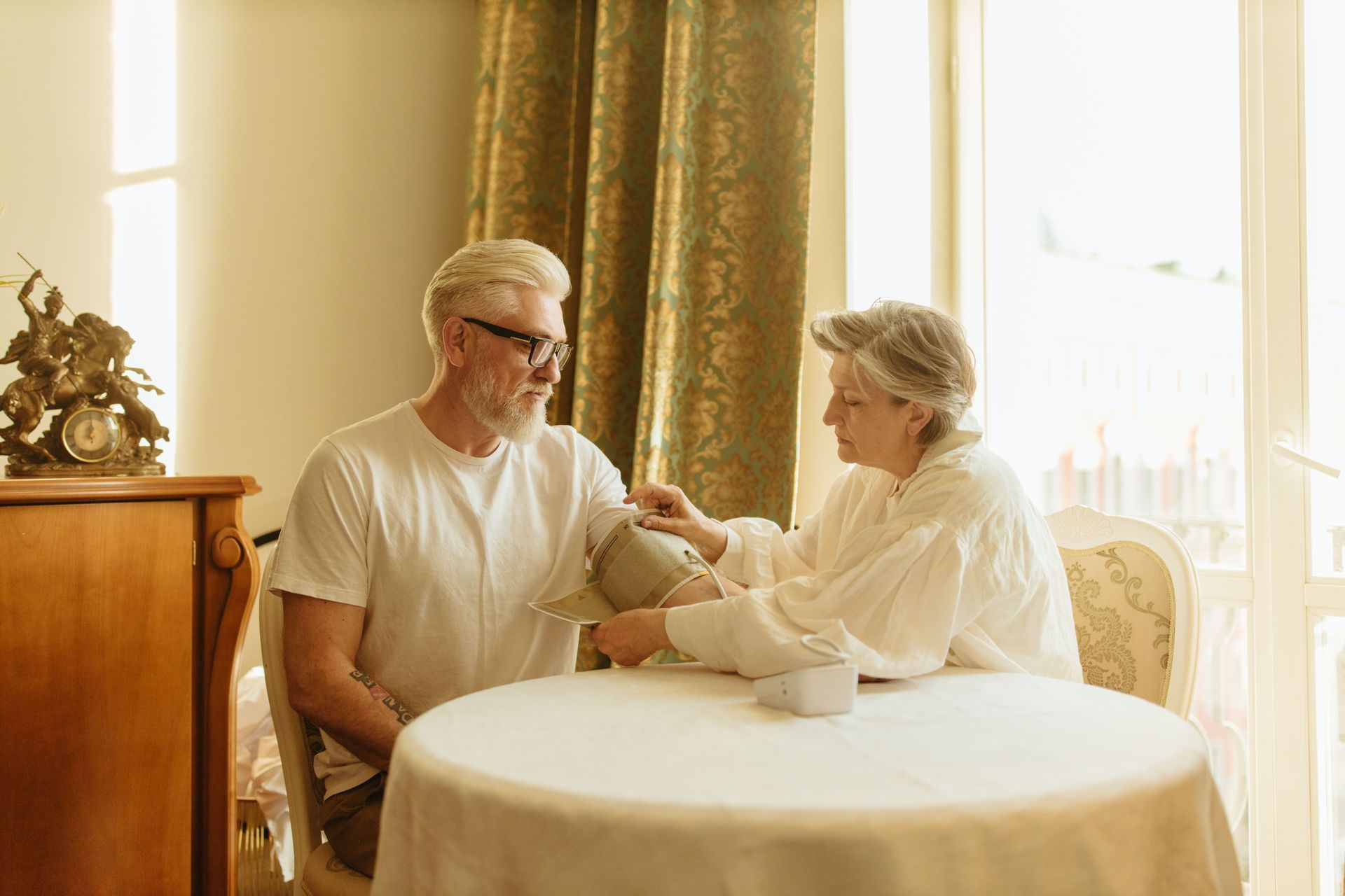 Two people sit at a sunlit table, talking and holding hands in a cozy room.