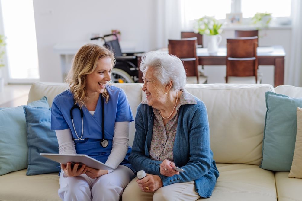 Two women talking on a couch, one in blue scrubs holding a tablet, in a bright living room