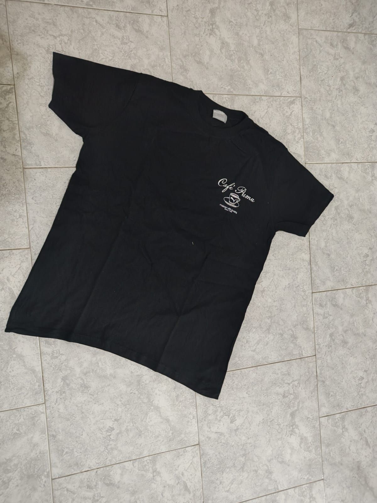 Camiseta negra con gráfico blanco en el pecho, colocada sobre un suelo de baldosas.