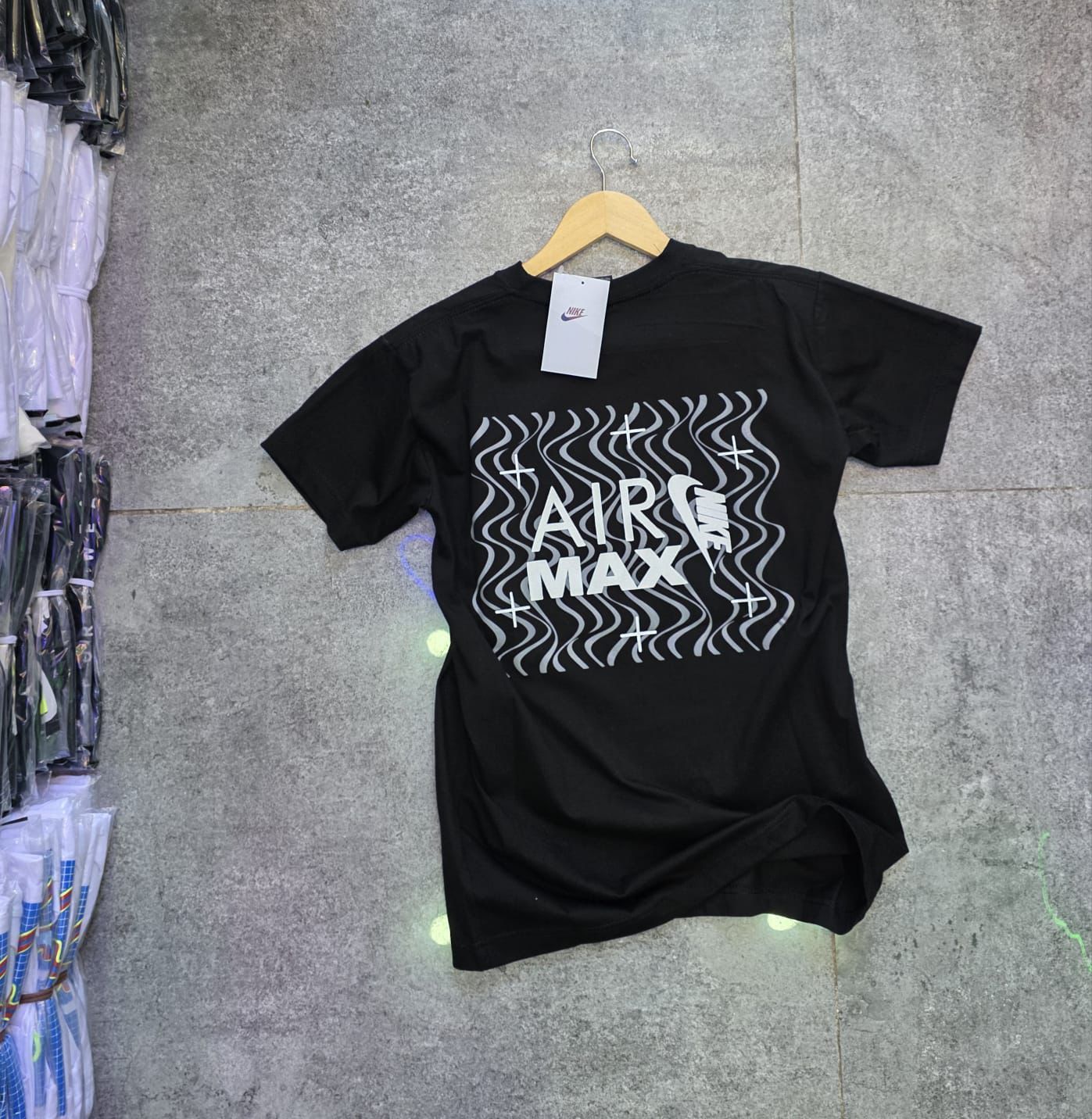 Camiseta negra Nike Air Max en una percha sobre una superficie gris. La camiseta tiene un gráfico blanco ondulado.