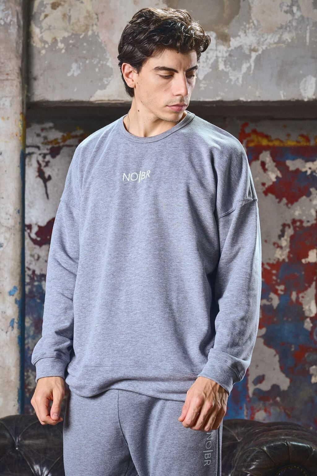 Un hombre está sentado en el suelo vistiendo una sudadera gris y pantalones deportivos.