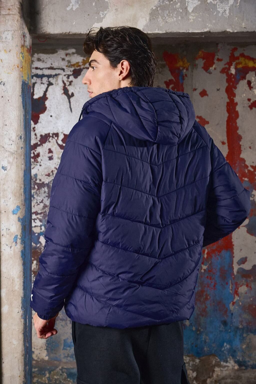 Un joven que lleva una chaqueta azul con capucha está parado frente a una pared.