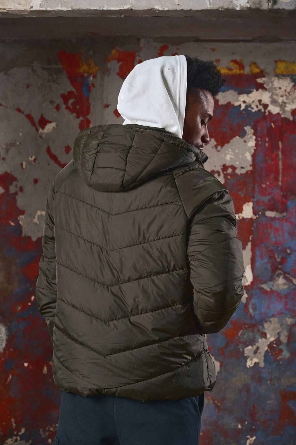 Un hombre que lleva una chaqueta verde y una sudadera con capucha blanca está parado frente a una pared.
