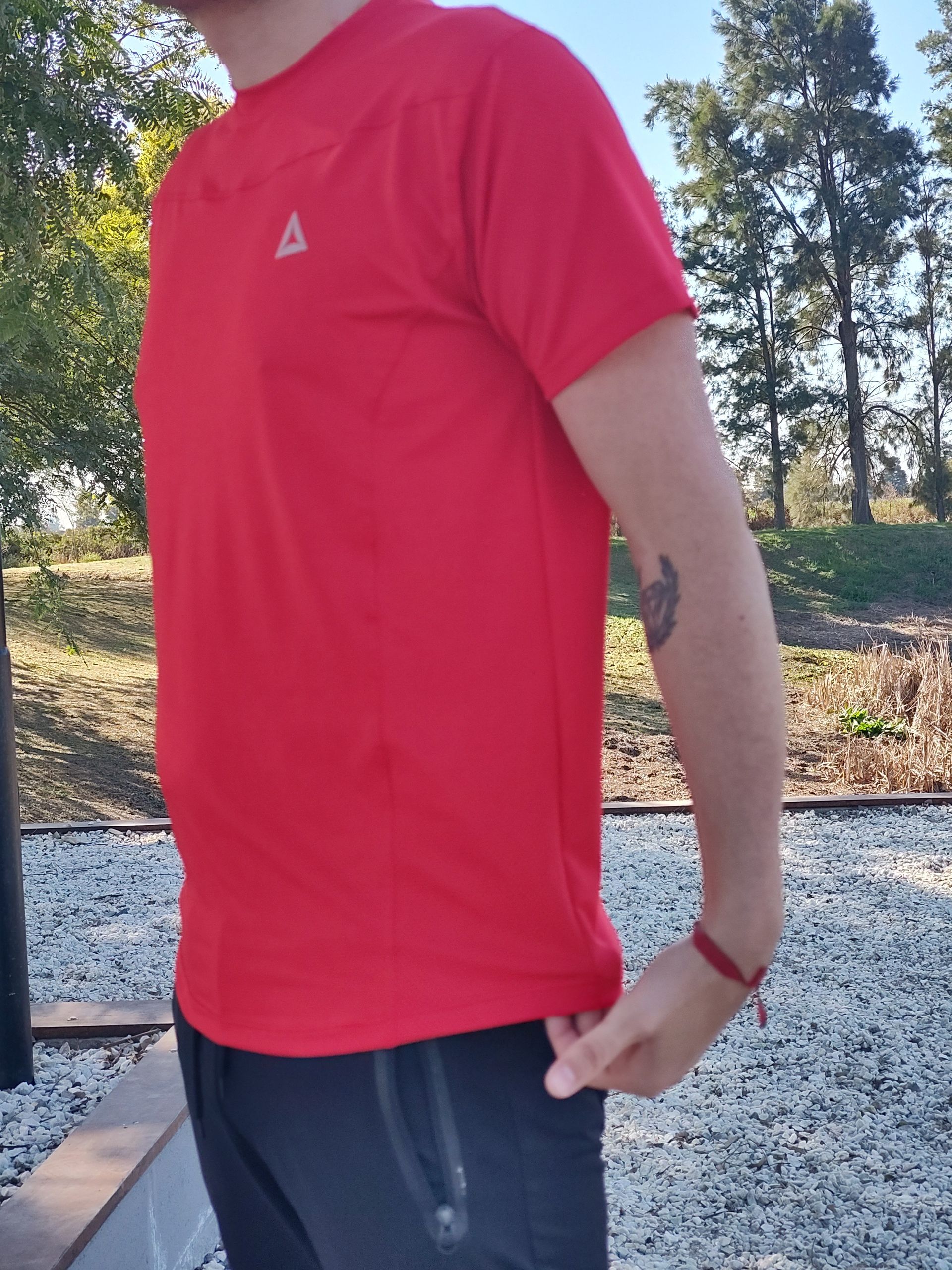 Un hombre con una camisa roja y las manos en los bolsillos sonríe, de pie al aire libre bajo un árbol.