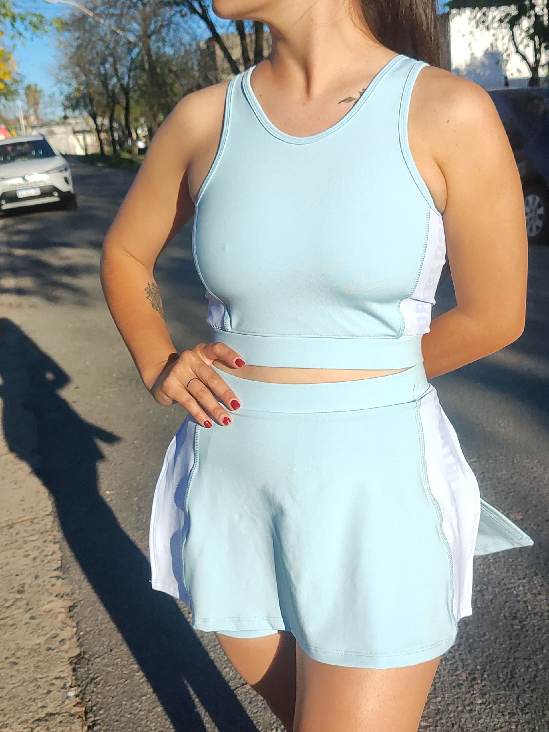 Mujer vestida con ropa deportiva azul claro, de pie al aire libre, con la mano en la cadera.