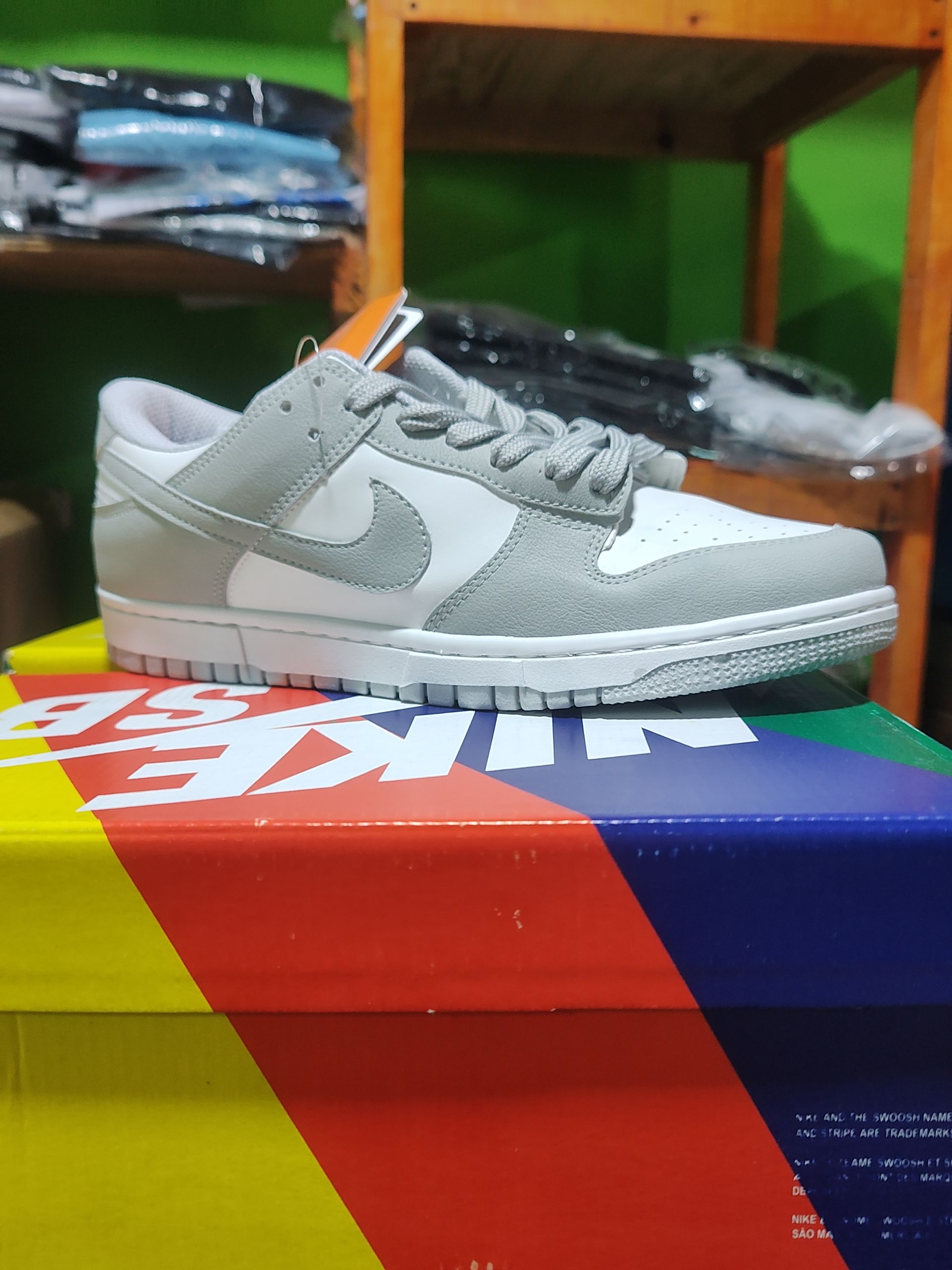 Zapatilla Nike Dunk Low gris y blanca en una caja colorida, con un estante de madera al fondo.