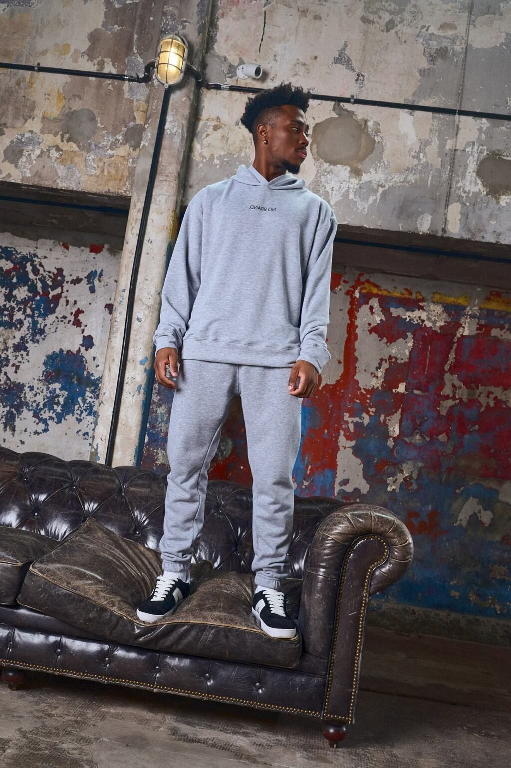 Un hombre con una sudadera con capucha gris y pantalones deportivos está de pie en un sofá.