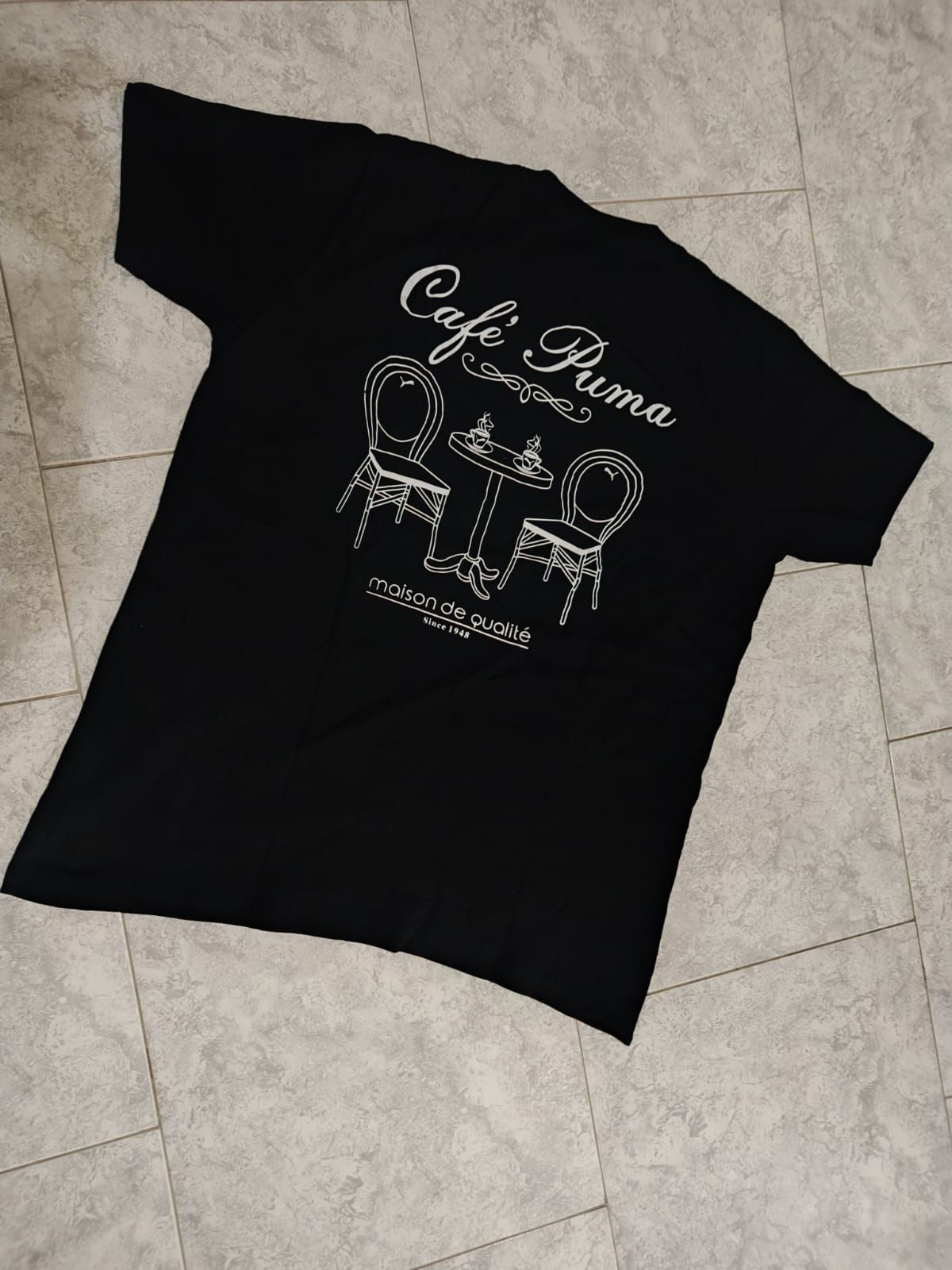 Camiseta negra con gráfico blanco en el pecho, colocada sobre un suelo de baldosas.