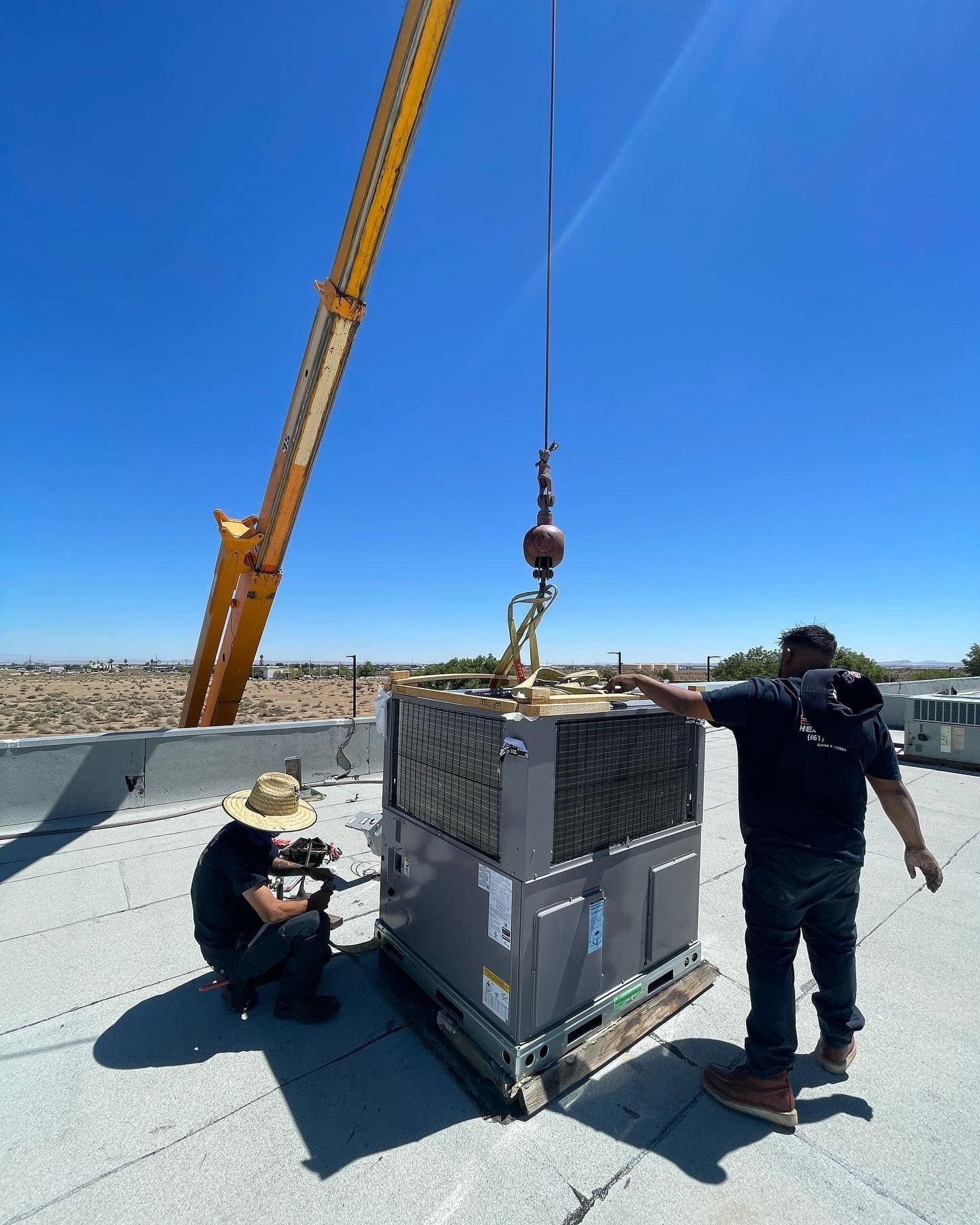 Two men are working on an air conditioner on a roof with a crane.