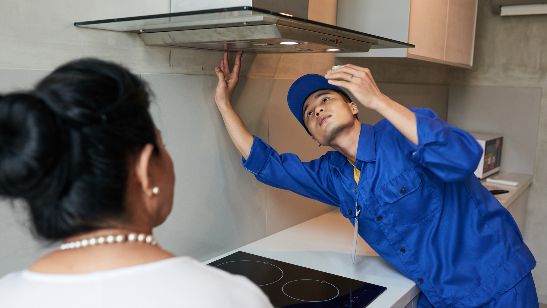 A man in a blue uniform is fixing a stove hood in a kitchen.