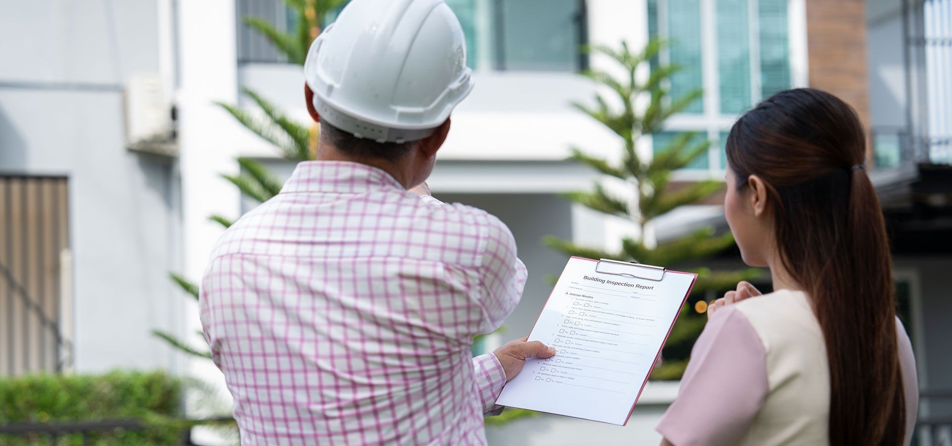 A man wearing a hard hat is standing next to a woman holding a clipboard.