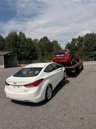 White Hyundai Elantra car parked next to a tow truck carrying a red SUV on a paved lot.