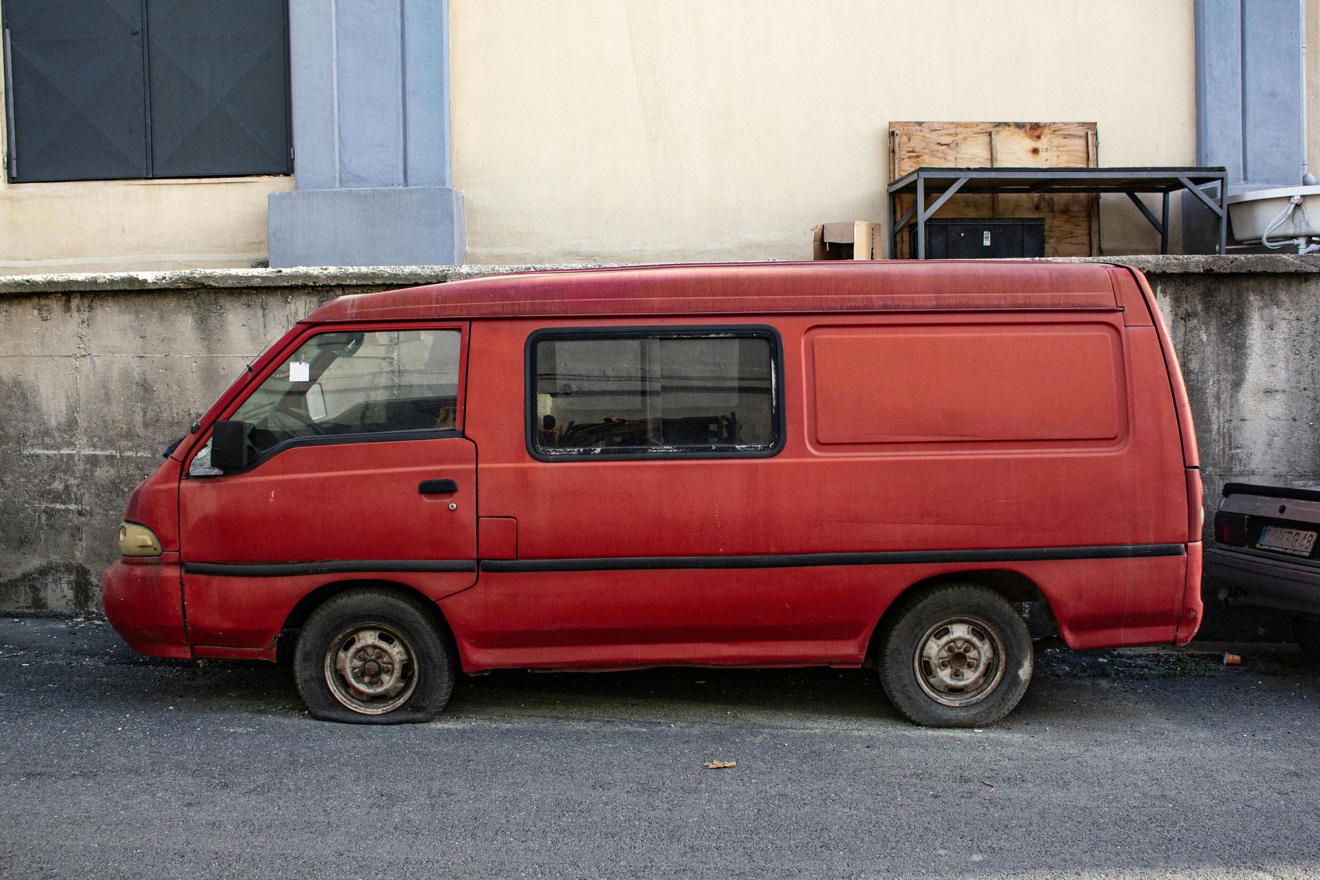 Red, weathered van parked on a gray street with a flat tire; building in the background.