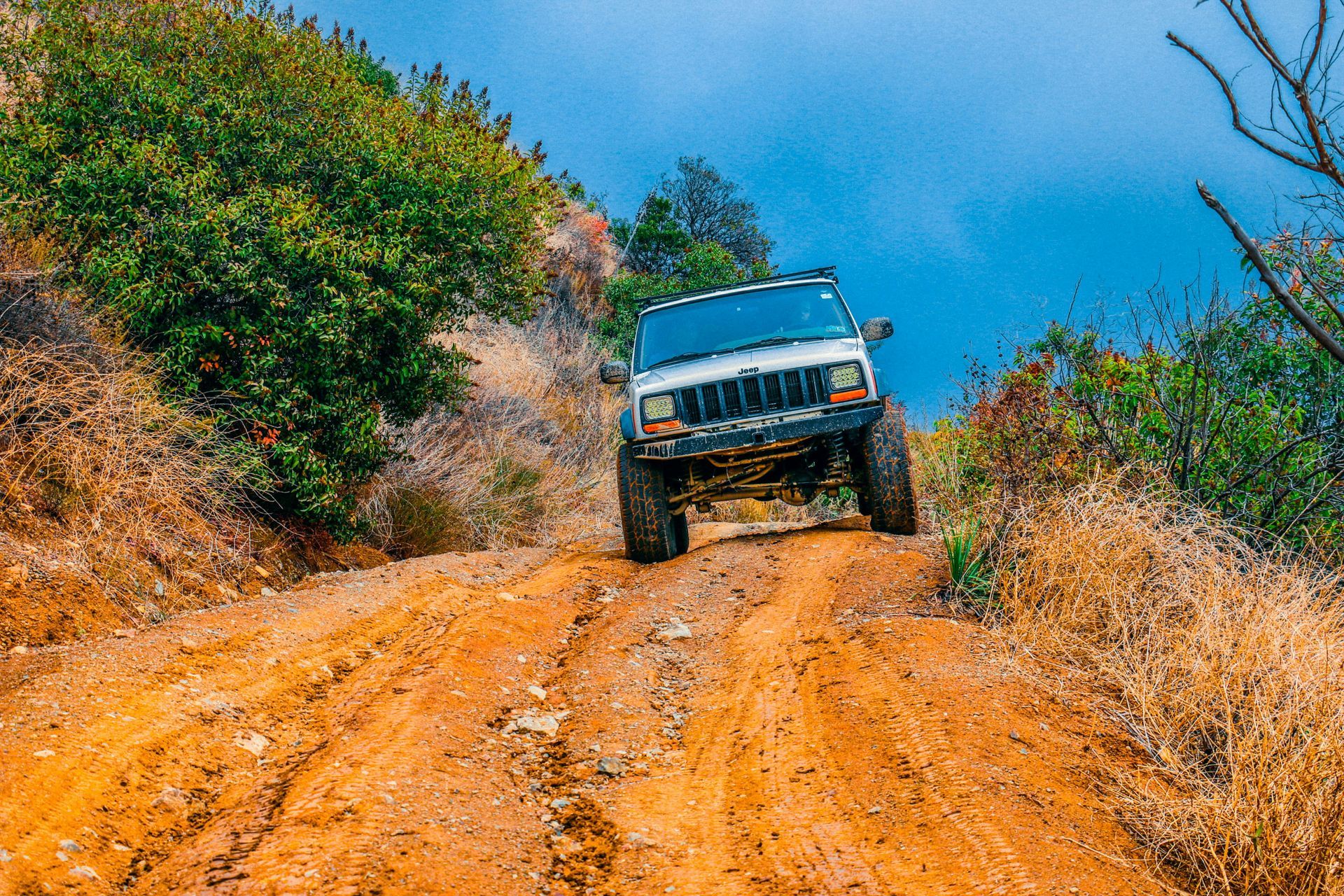 White Jeep climbs a steep, dirt hill. Overcast sky, brown and green vegetation surrounds.