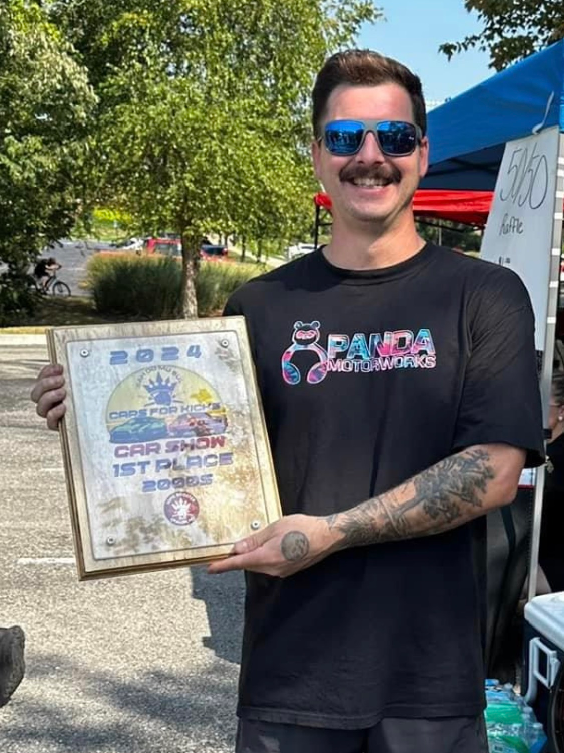 A man wearing sunglasses and a mustache is holding a framed certificate.