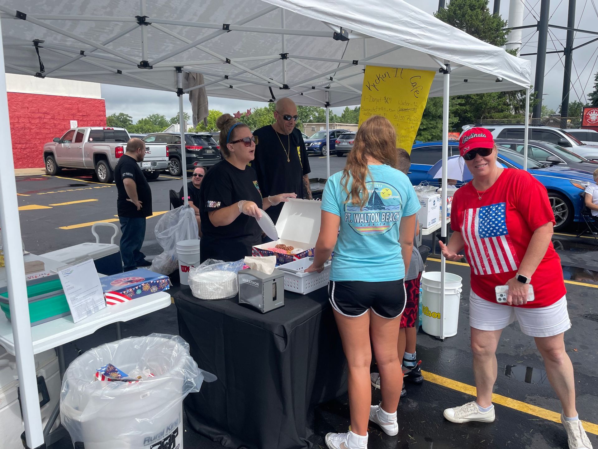 A group of people are standing around a table under a tent in a parking lot.