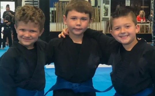 Three young boys wearing black karate uniforms and blue belts are posing for a picture.