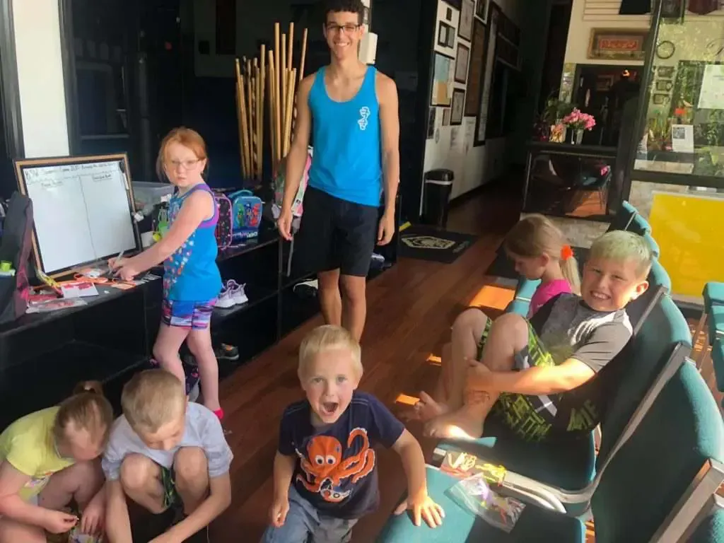 A group of children are sitting in a waiting room with a man standing behind them.