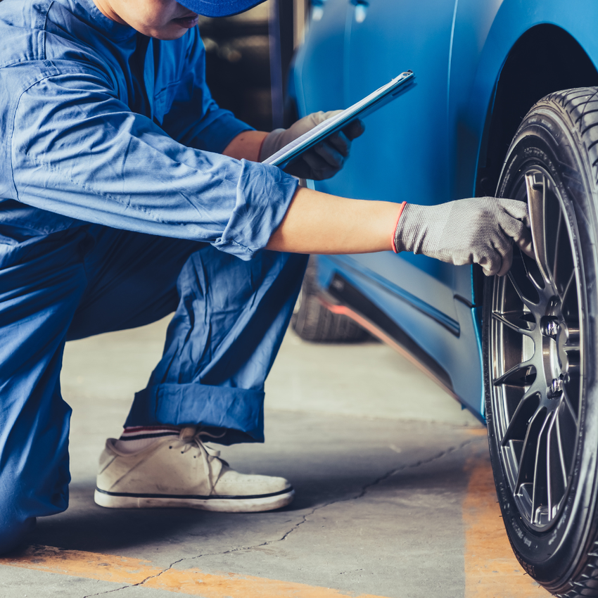 Mechanic crouching beside a blue car, inspecting the wheel with a tablet and glove on