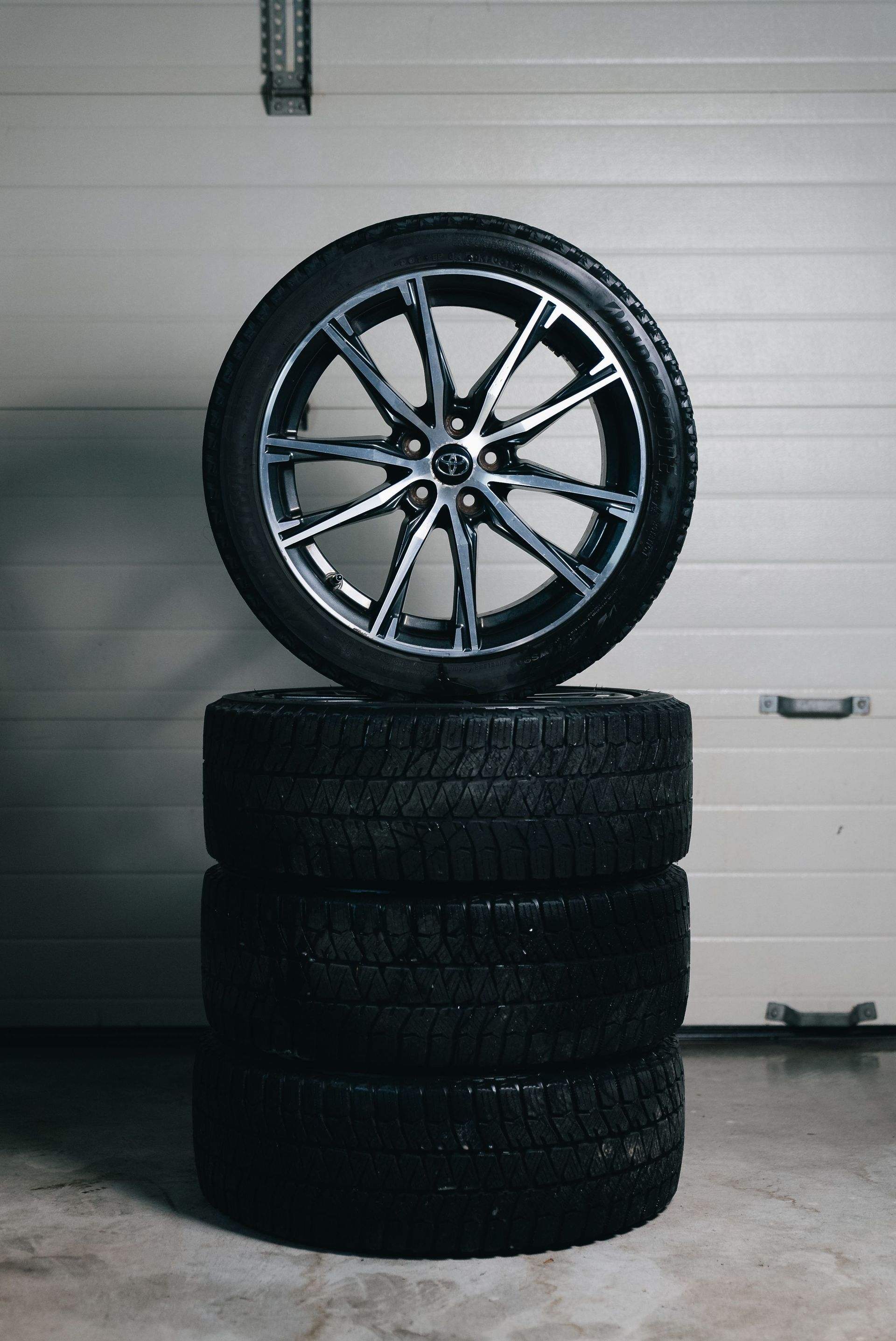 Car wheel rim balanced on four stacked black tires in a garage.