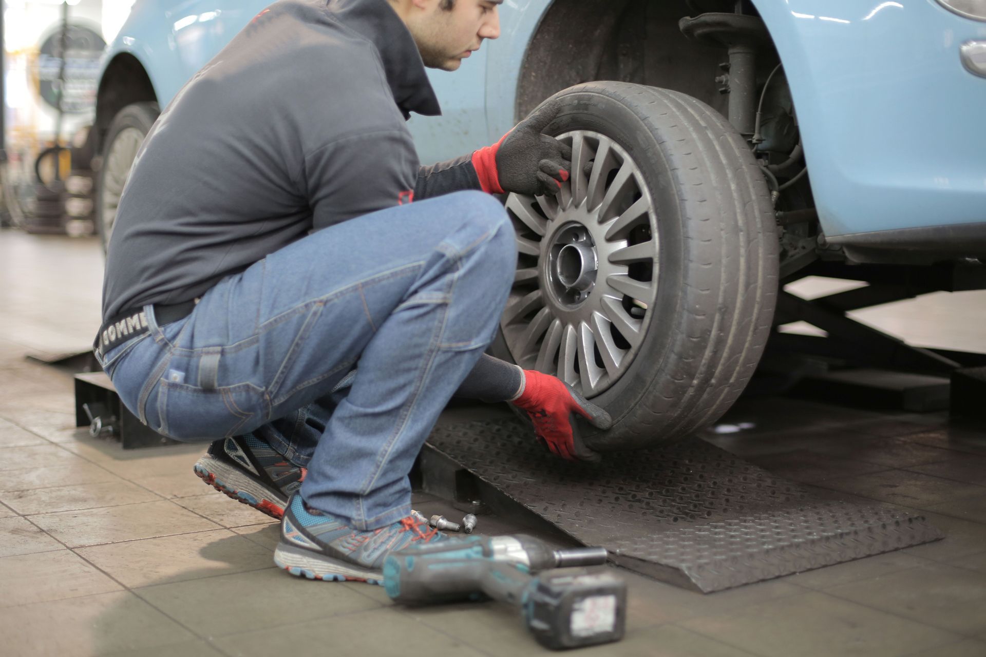 Mechanic kneeling beside a blue car, inspecting the wheel with a tire tool in a garage