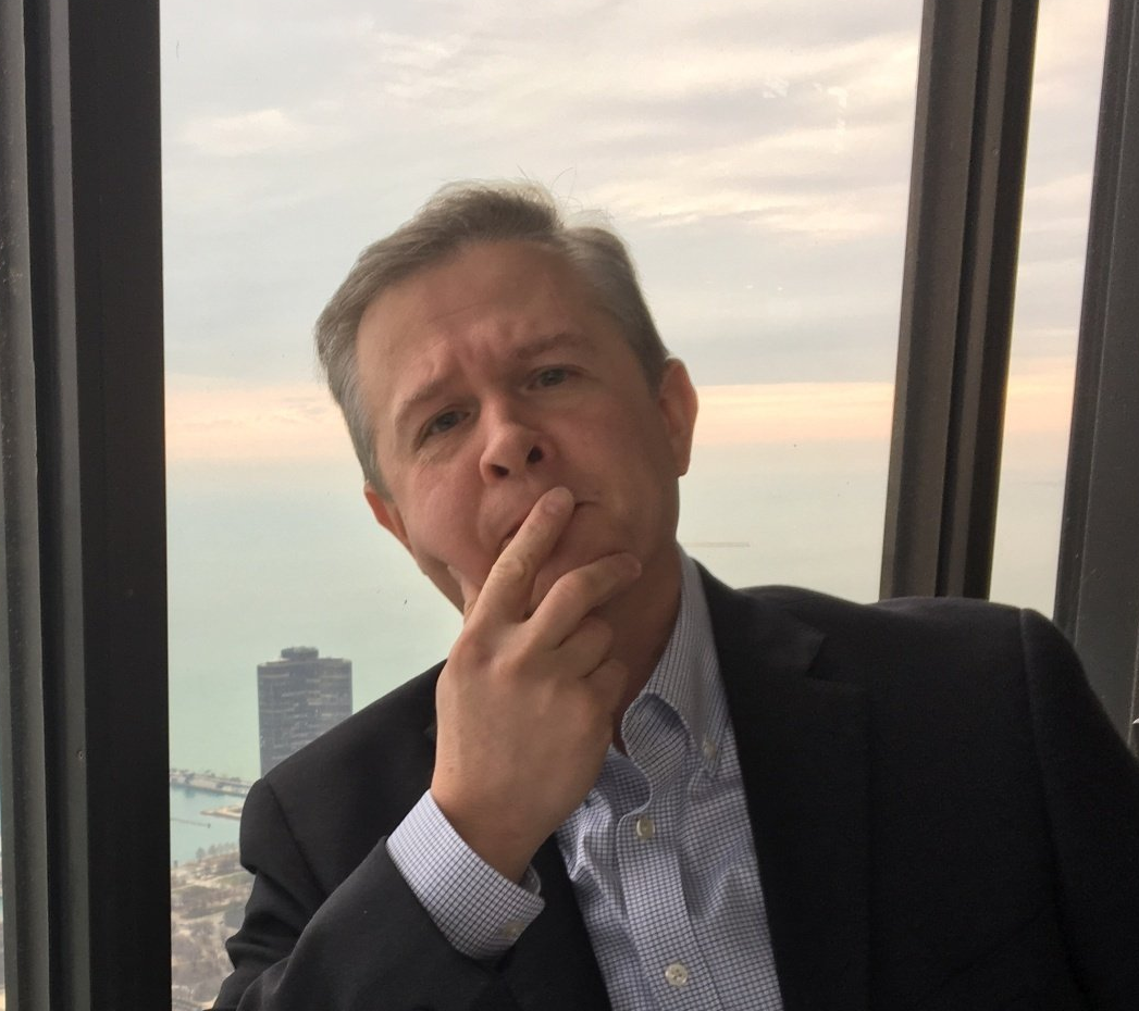 A man in a suit is sitting in front of a window with his hand on his chin