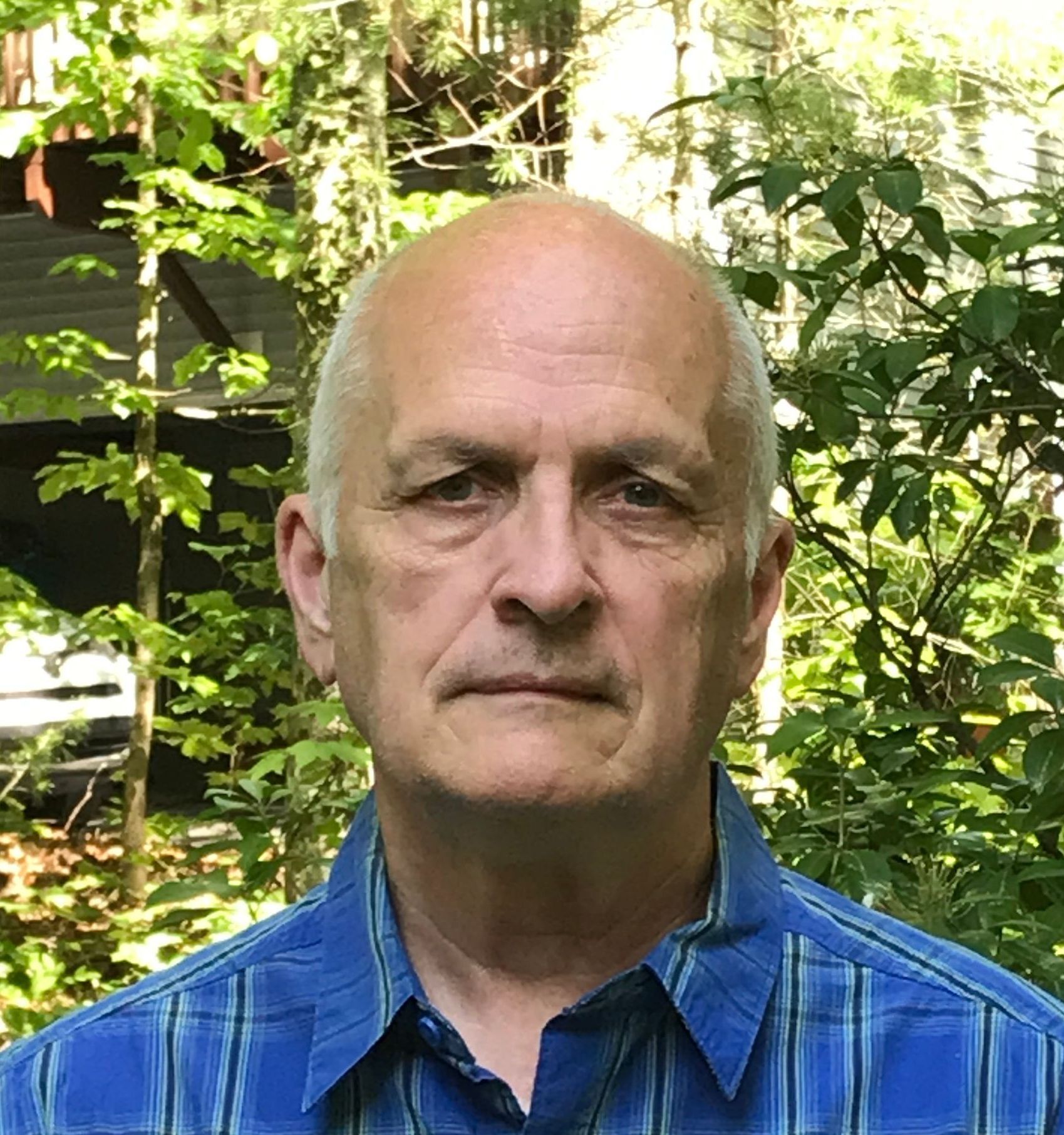 A bald man in a blue plaid shirt is standing in front of trees.