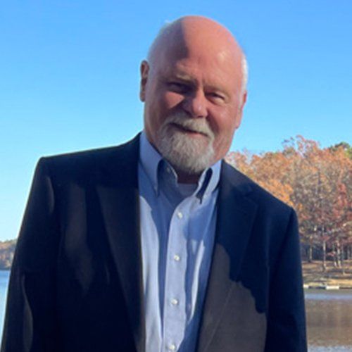 A man in a suit and blue shirt is standing in front of a lake.