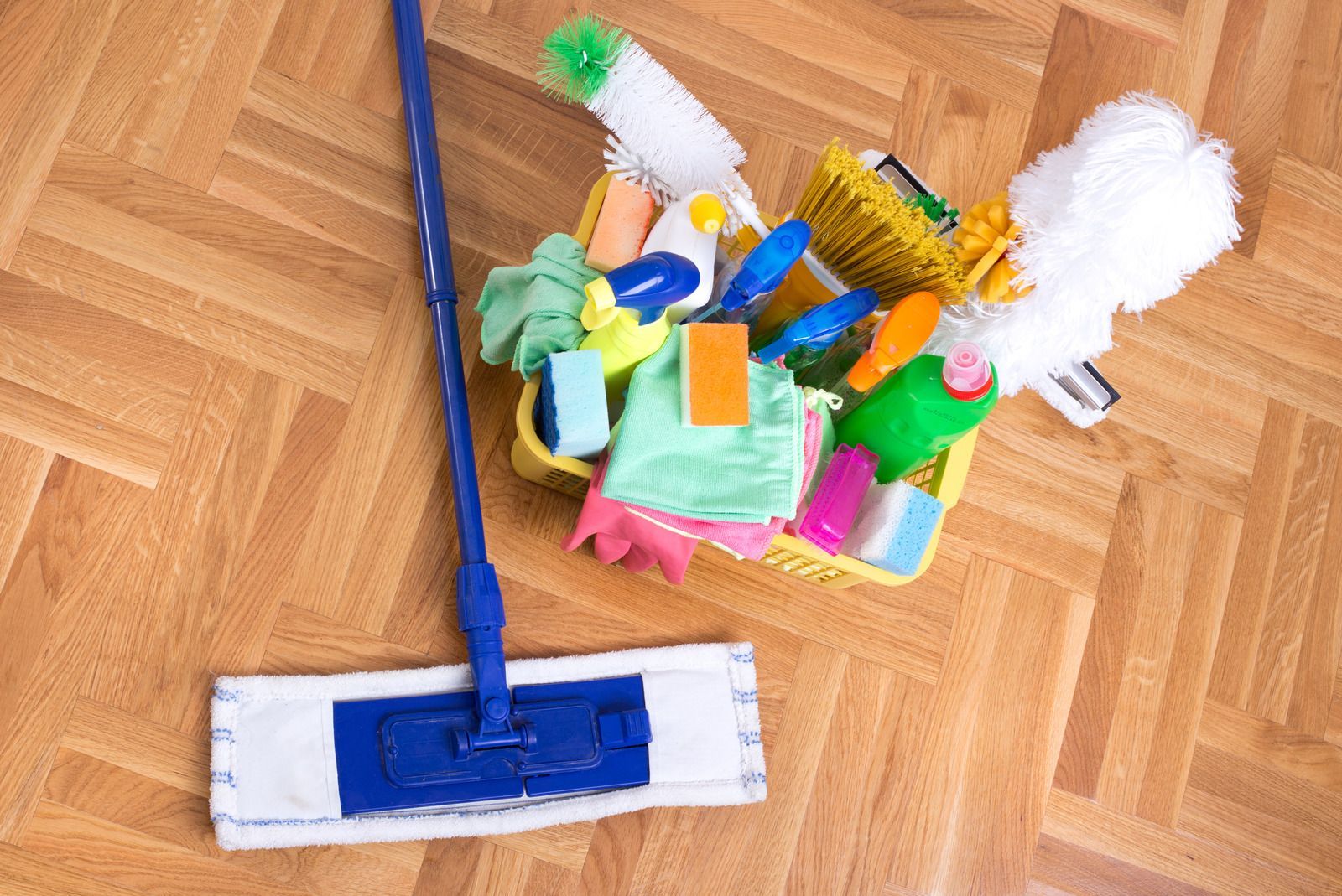 A mop is sitting on a wooden floor next to a basket of cleaning supplies.