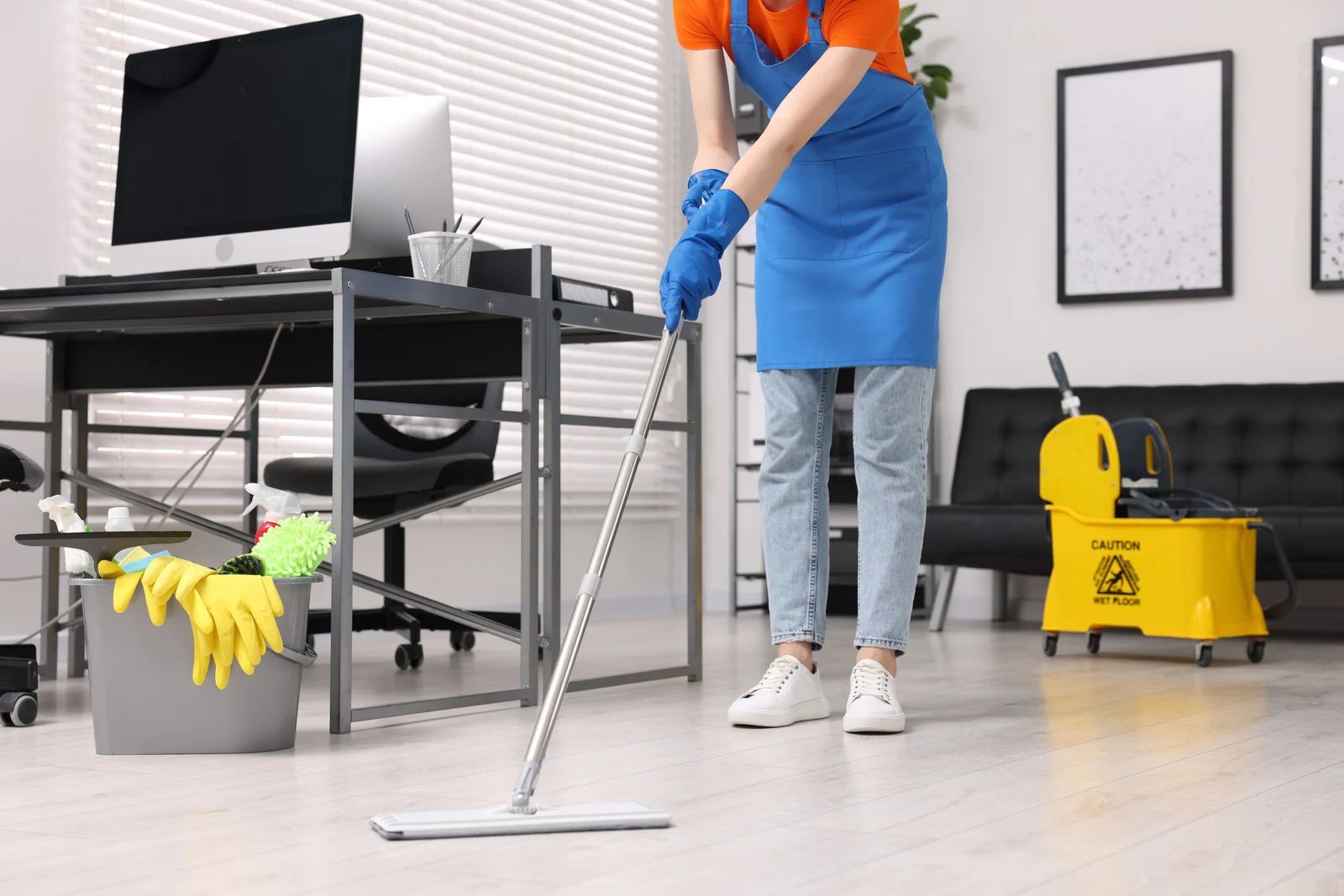 Person mopping an office floor with a gray mop. They wear blue gloves, apron, and jeans.