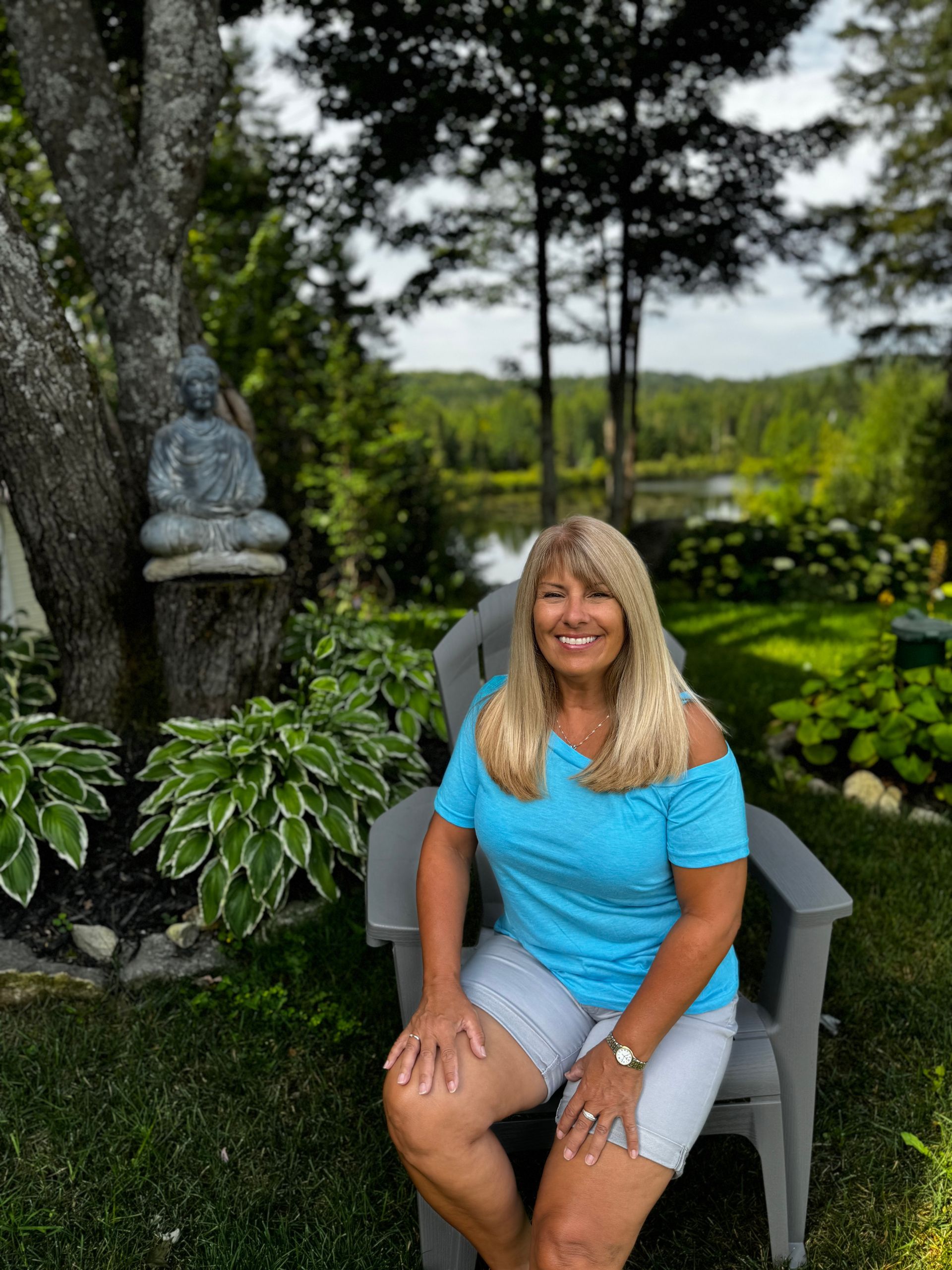 Femme en haut bleu et short blanc assise sur une chaise de jardin, avec une statue de Bouddha en arrière-plan.