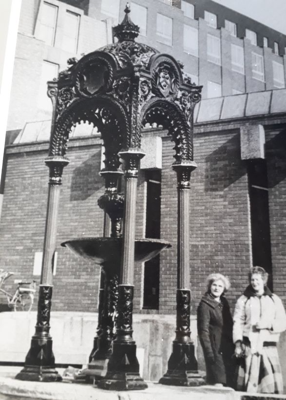 Black and white photo of ornate fountain and two women standing beside it. Brick building in the background.