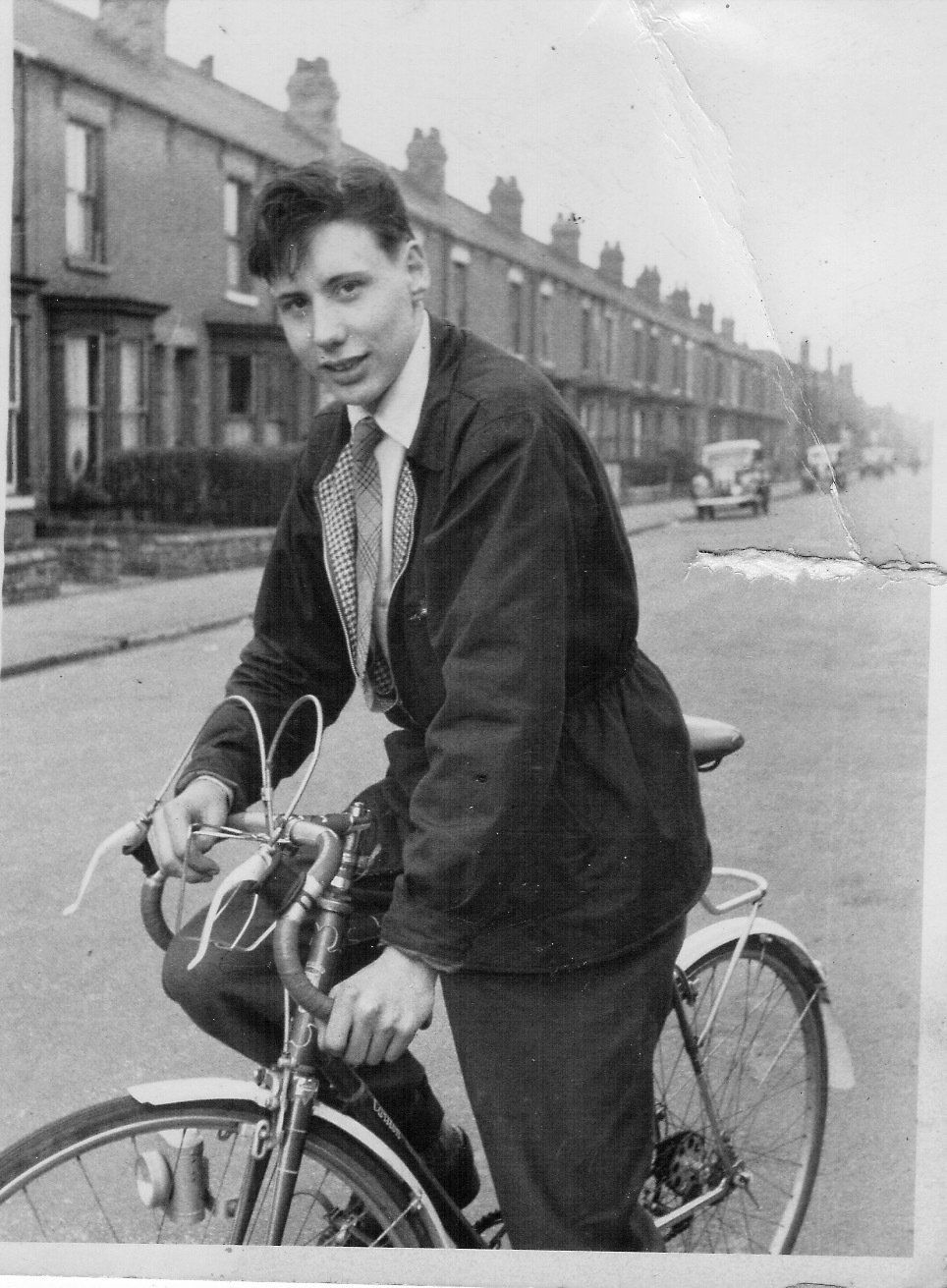 Young man on a bicycle on a street. He wears a jacket, tie, and pants. Houses line the street.