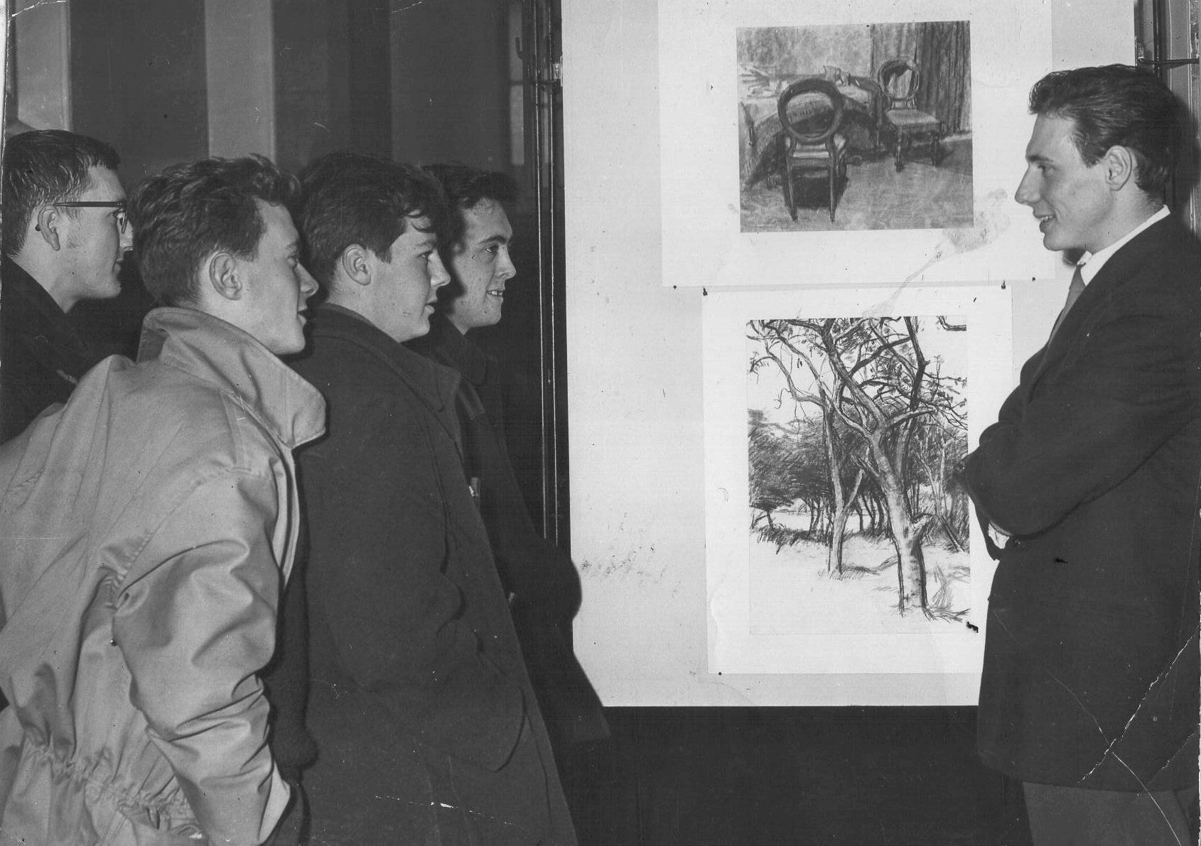 Group of men looking at artwork displayed on a wall. One man gestures towards the art while others observe.